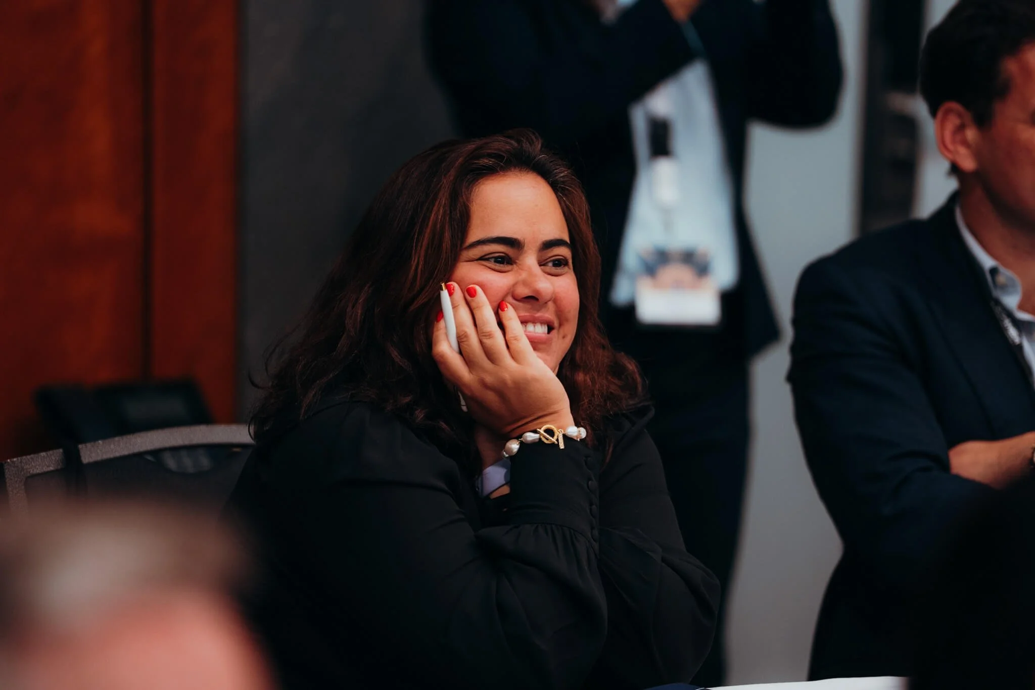A woman with dark brown hair smiling and resting her face on her hand during a meeting or conference.
