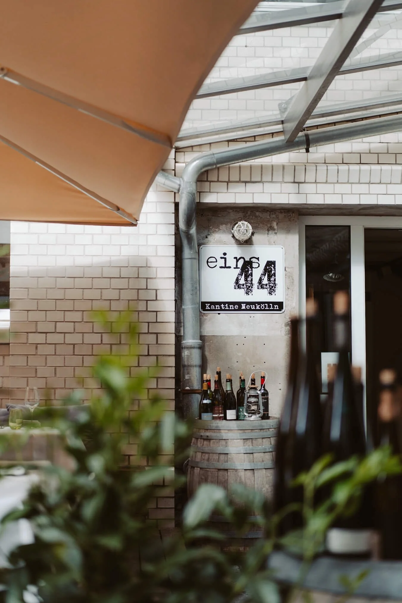 Outdoor seating area of a restaurant or café with a sign that reads 'eins 44 Kantine Neukölln,' a barrel with bottles on top, and greenery in the foreground.