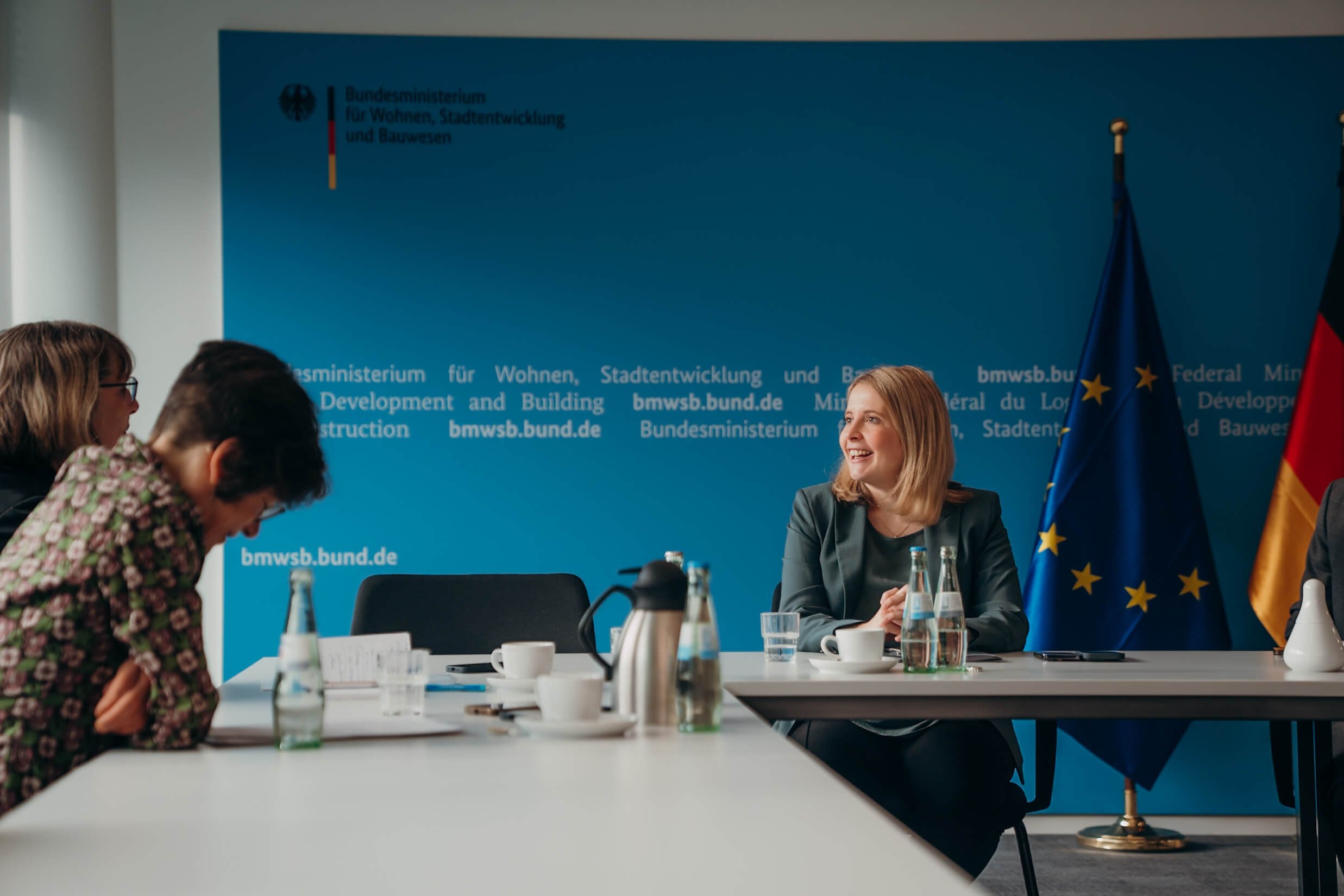 Eine Frau mit blonde Haare sitzt bei einer offiziellen Besprechung, im Hintergrund eine blaue Wand mit dem Logo des Bundesministeriums für Wohnen, Stadtentwicklung und Bauwesen, sowie der deutschen und europäischen Flagge.