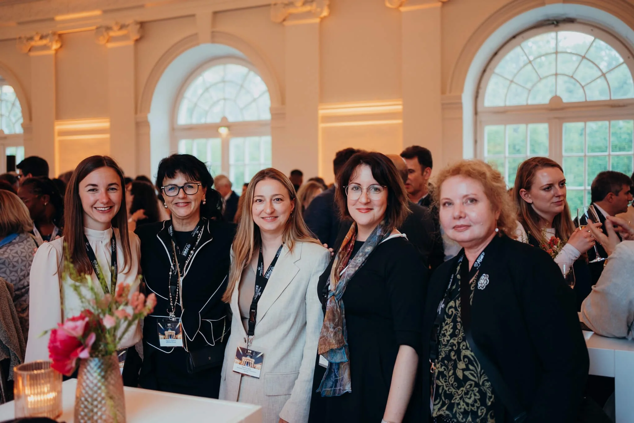 Five women standing together at a formal event in a large, well-lit room with arched windows and many attendees in the background.