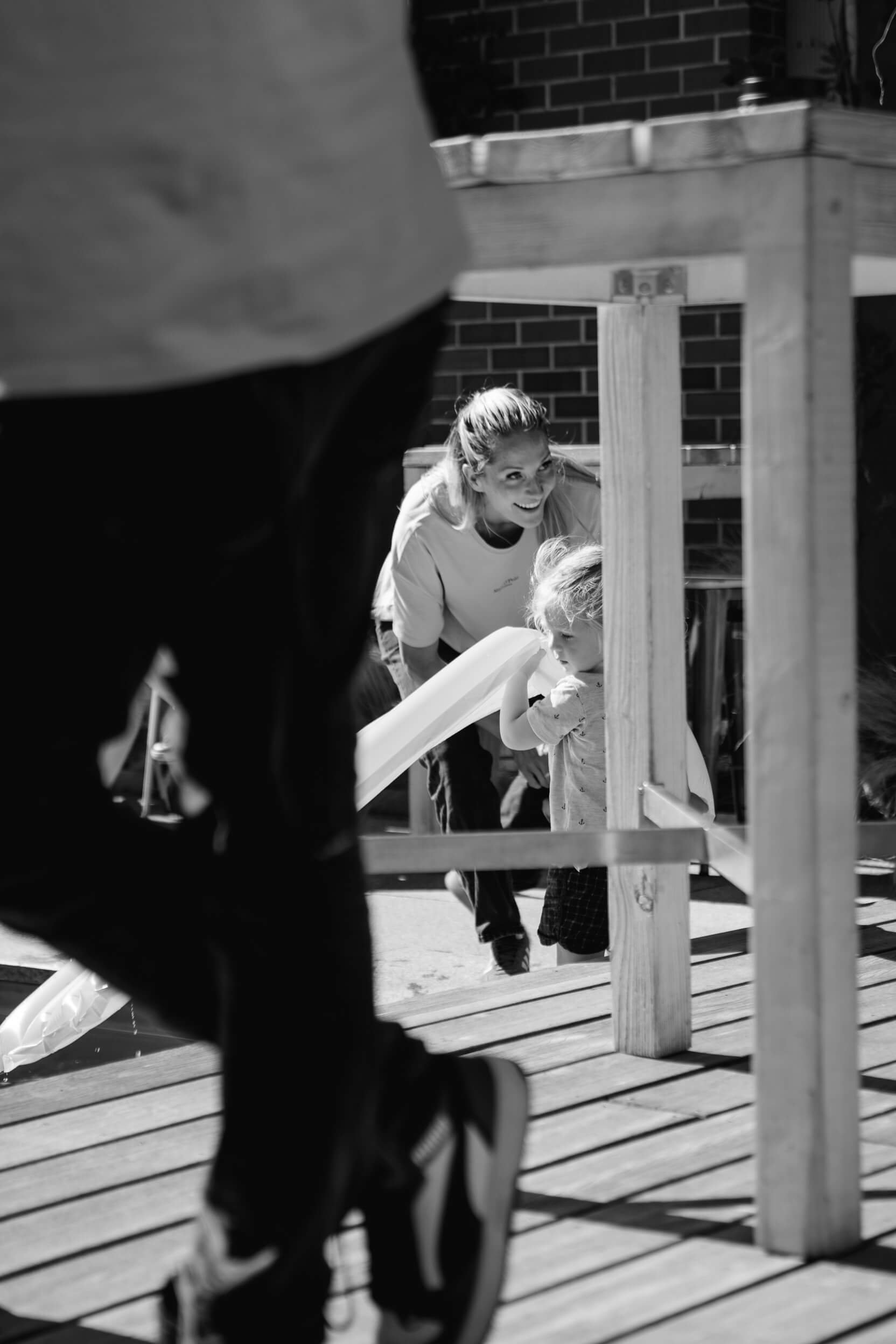 Ein Mädchen und ein Kleinkind spielen auf einem Holzspielplatz. Das Mädchen lächelt, während das Kind in der Nähe steht. Im Vordergrund sind Schuhe zu sehen, teilweise durch eine Person verdeckt.