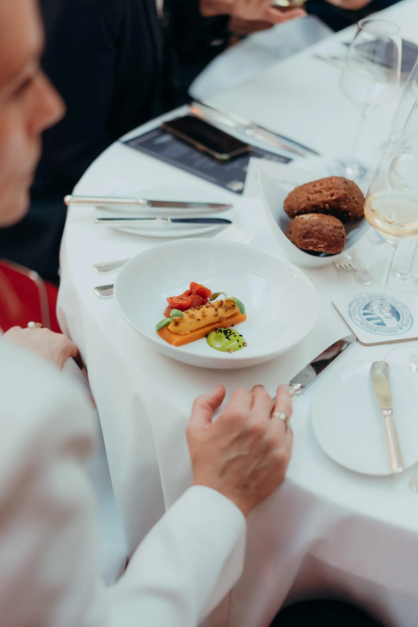 People dining at a formal event, with plates of food, wine glasses, and cutlery on a white tablecloth.
