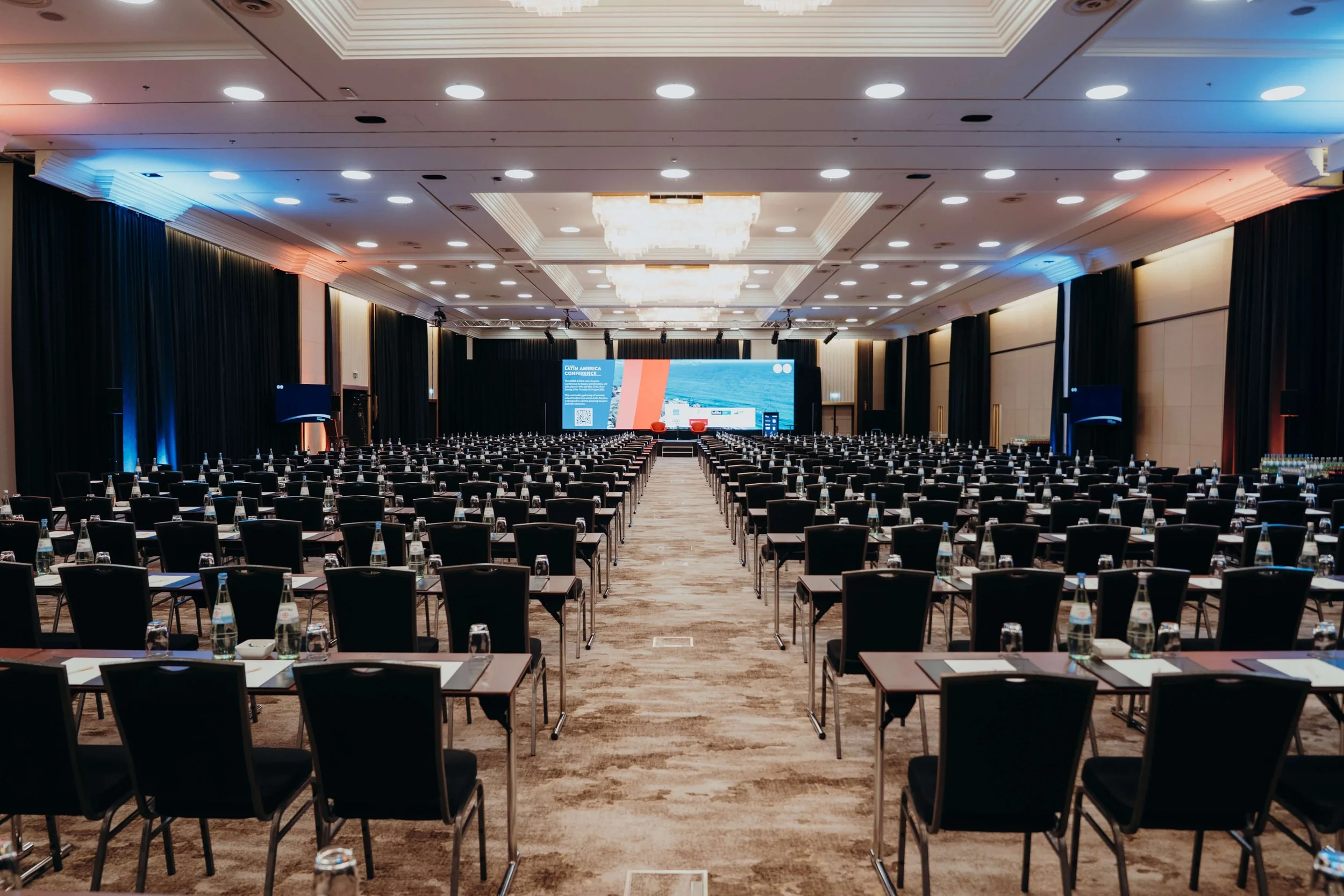 Empty conference room with rows of black chairs and tables, set with water bottles and notepads, facing a large stage with a big digital screen displaying conference information.