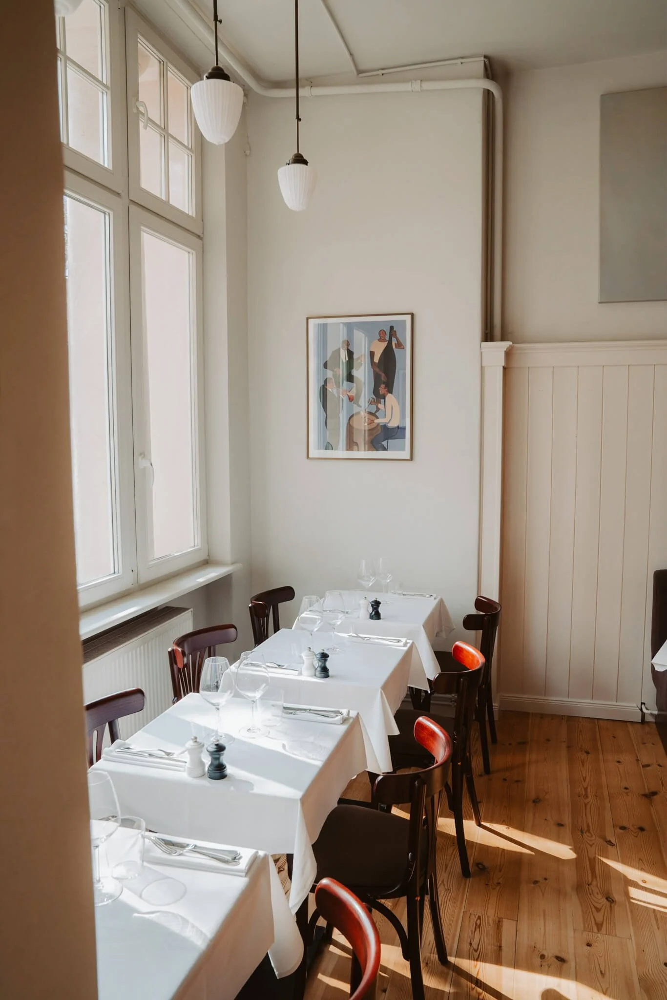 A small dining area with four tables covered in white tablecloths, set with wine glasses, silverware, and small salt and pepper shakers. The room has large windows letting in natural light, white walls, wooden floors, and a framed artwork on the wall