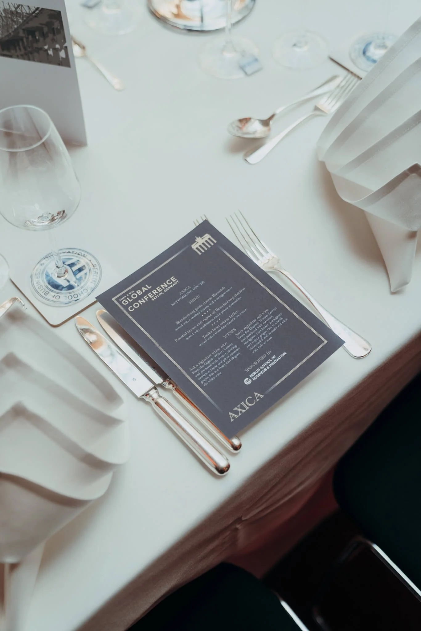 Table setting with silverware, wine glass, and a program/menu for an event, possibly a conference, with a glass and some other tableware in the background.