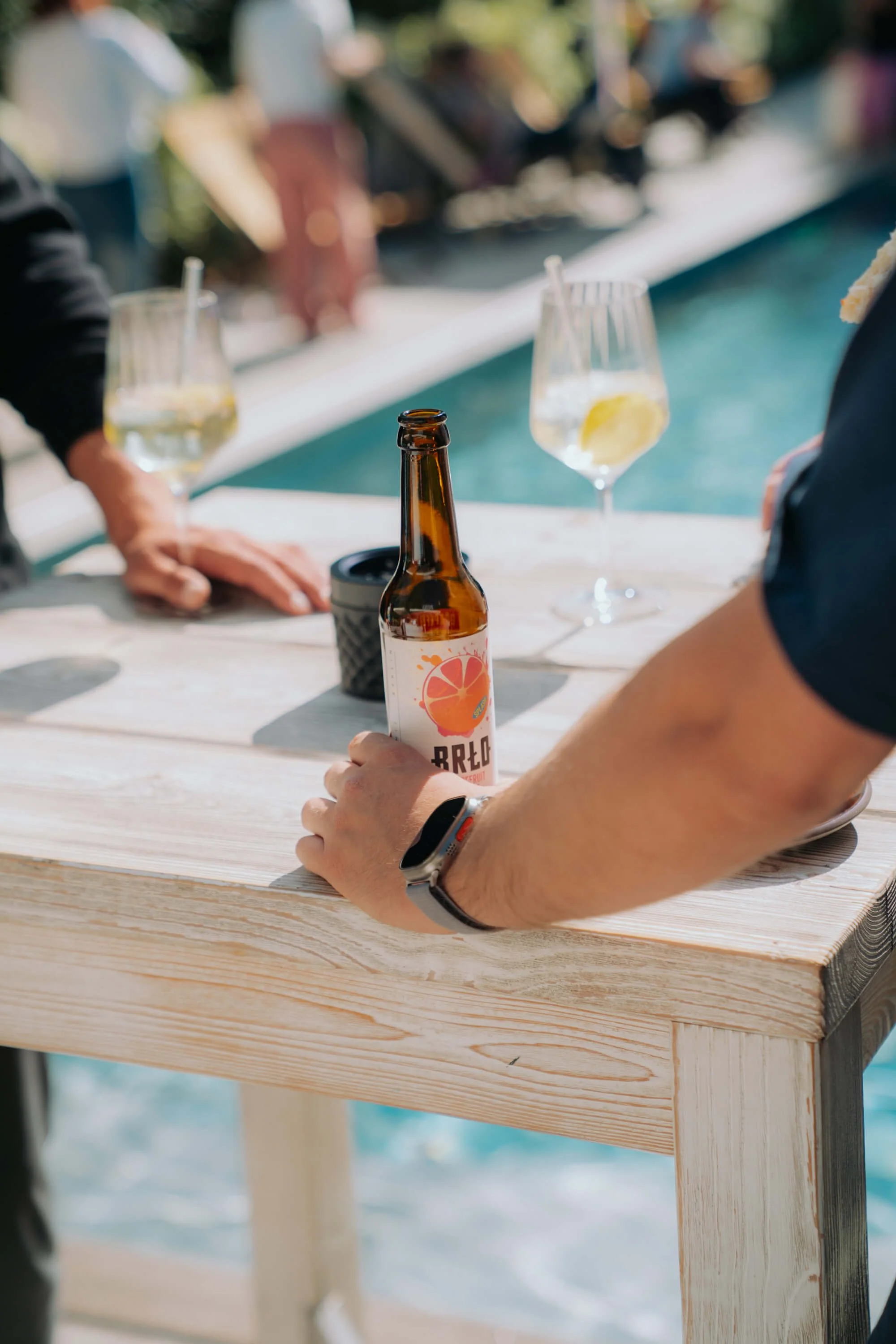Menschen, die an einem hellen Holztisch sitzen, trinken Getränke mit Zitronenscheiben in Weingläsern, während sie sich unterhalten, im Hintergrund ist ein Pool und Bäume, die Szene ist hell und sommerlich.