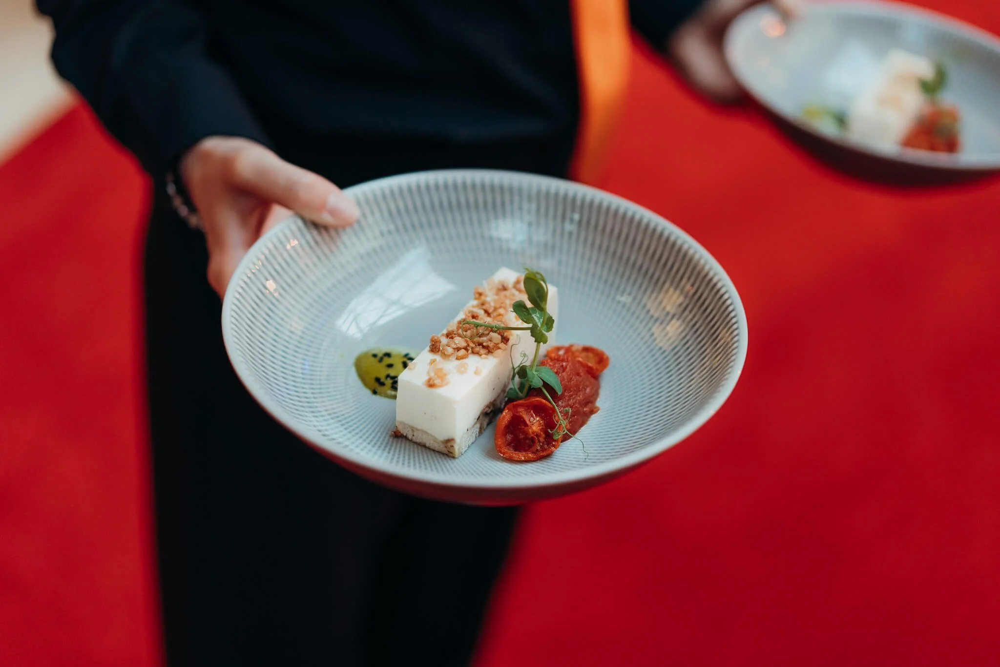 A person holding a bowl of gourmet dessert with a rectangular piece of white mousse garnished with crushed nuts and a small green herb, alongside a dollop of red sauce or fruit compote, on a red table.