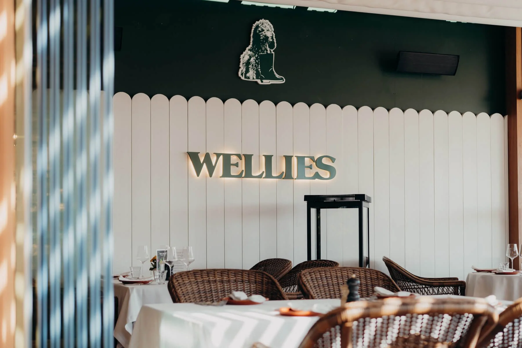 Ein Restaurant mit weißen Tischen und Stühlen, einer Wand mit dem Schriftzug 'WELLIES' und einer Wanddekoration eines Löwenkopfes mit Stiefel.