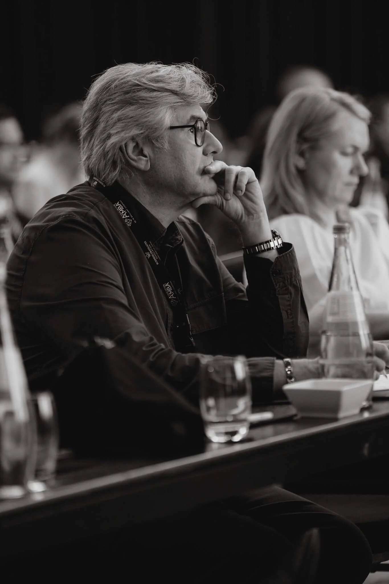 A man with glasses and gray hair, sitting at a conference table with a pen in his hand and a water bottle and glass in front of him, listening intently.