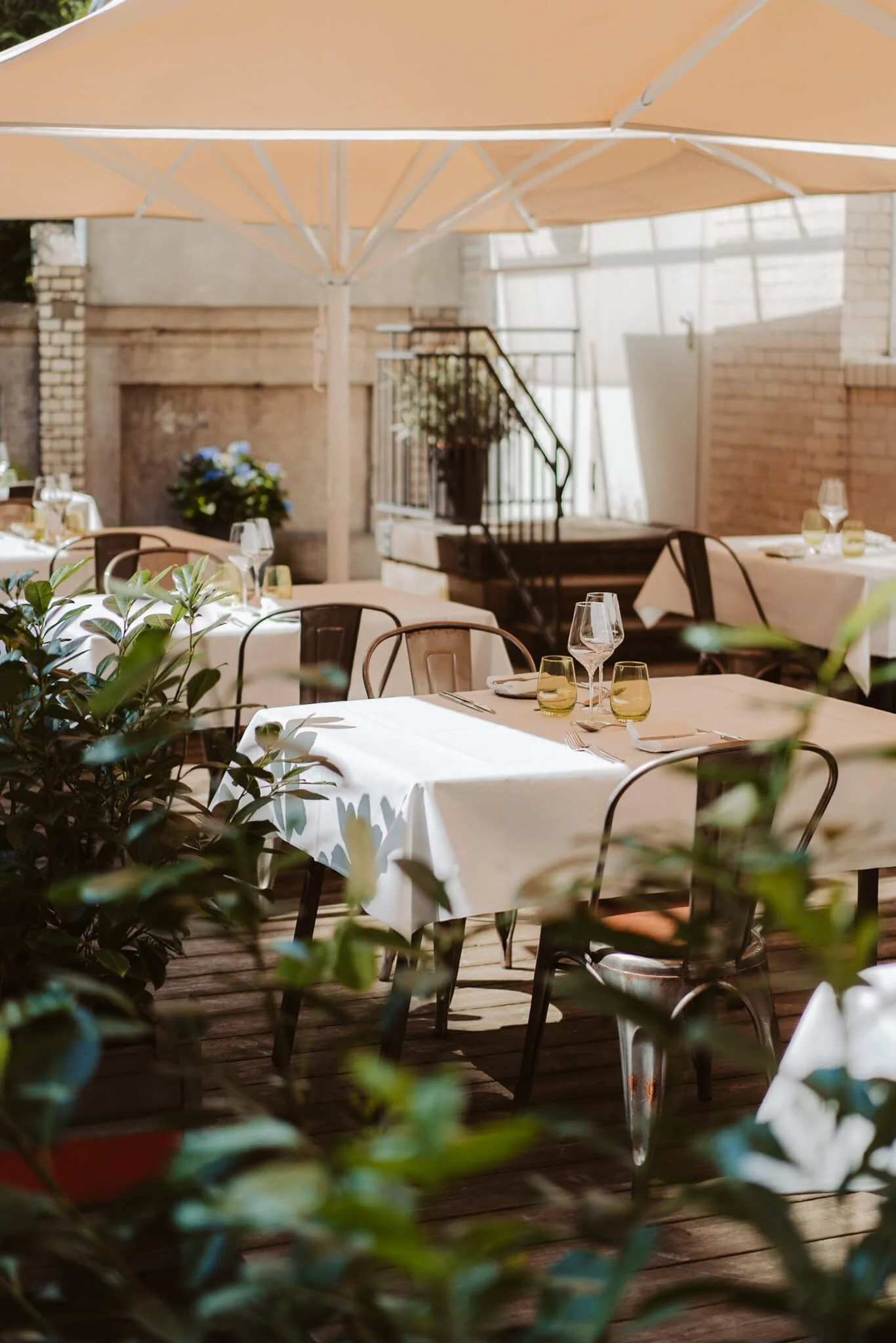 Outdoor restaurant patio with white tablecloth-covered tables, chairs, glassware, and potted plants, shaded by a large beige umbrella.