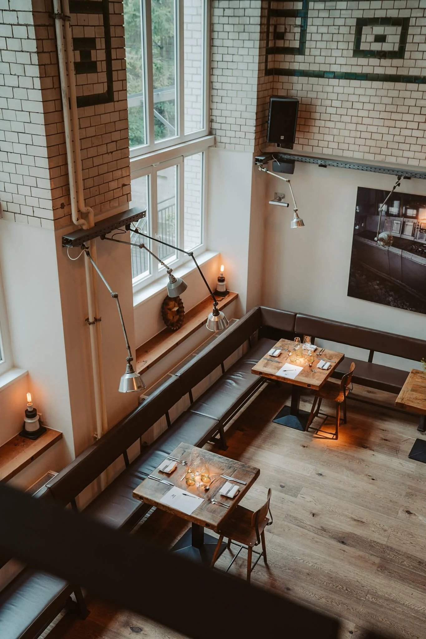 Interior of a cozy restaurant with wooden floors, a long leather banquette seating, and small tables set for dining, featuring napkins and glasses, illuminated by hanging pendant lights, with large windows letting in natural light, and industrial-sty