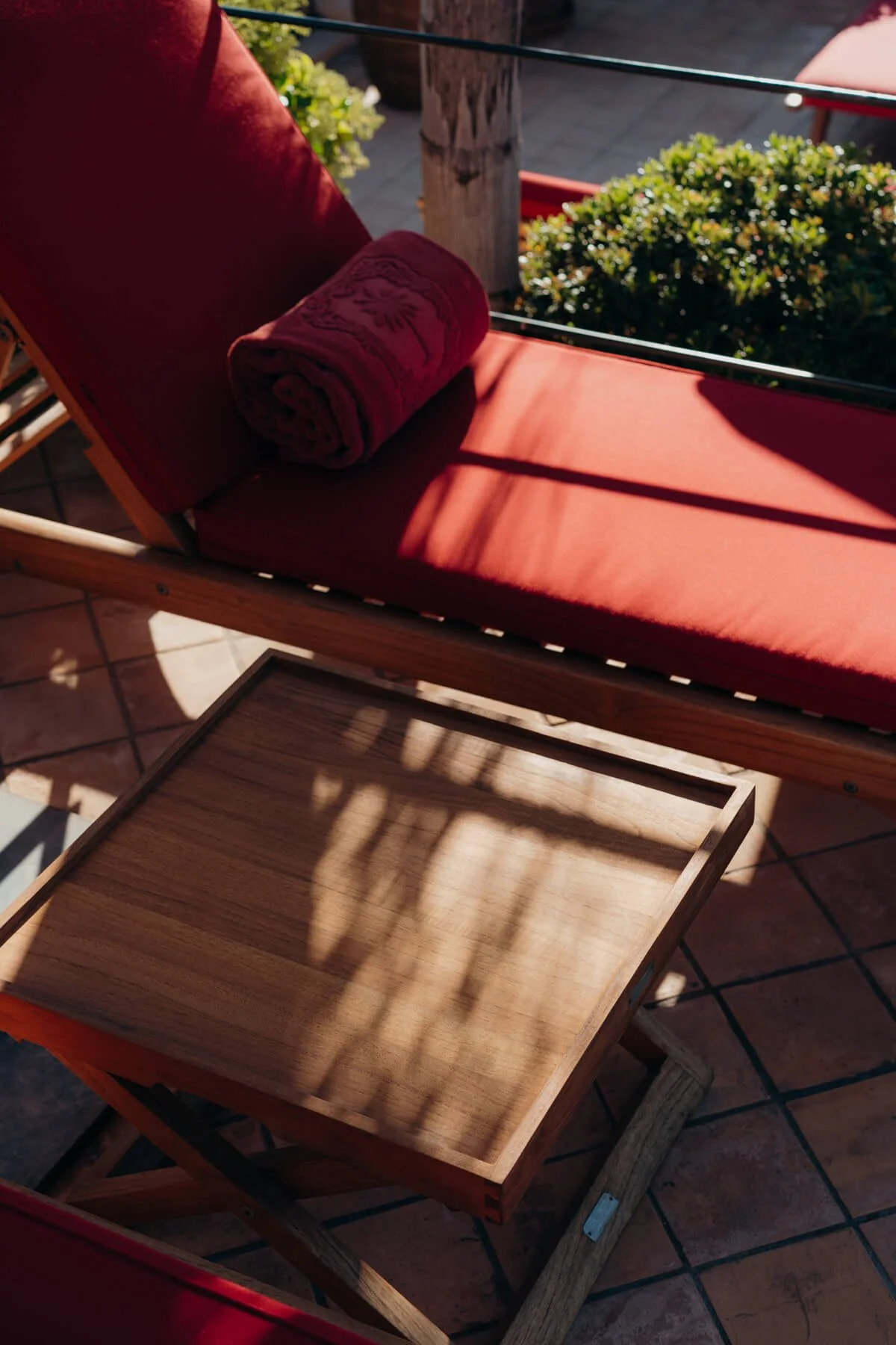 Ein rotes Liegestuhl mit einem im gleichen Farbton gewickelten Handtuch auf einer Terrasse mit Fliesenboden. Im Hintergrund sind grüne Büsche und ein Holzzaun zu sehen.