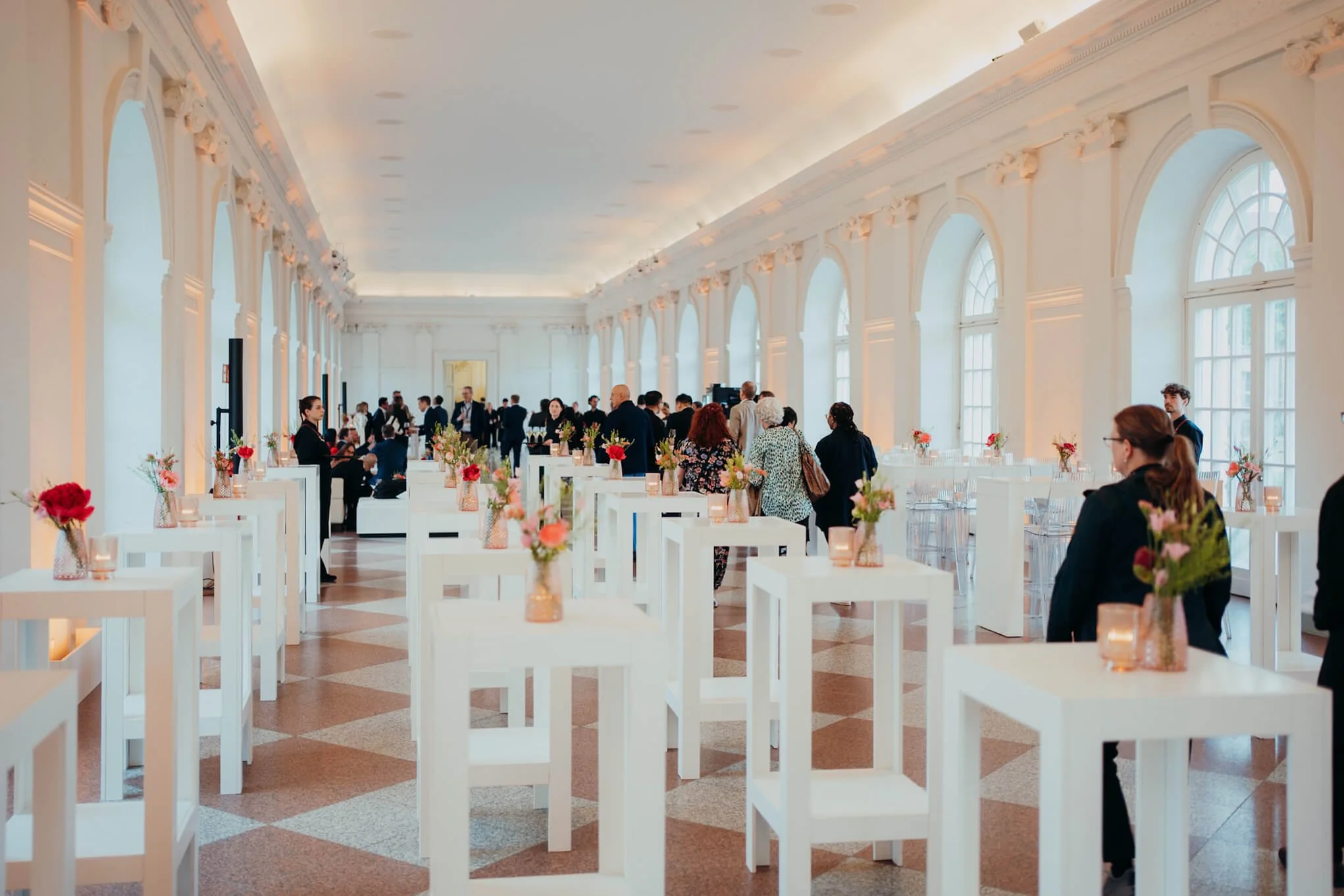 Elegant event space with tall arched windows, white walls, decorated with vases of pink and red flowers on white tables, people socializing and standing around.