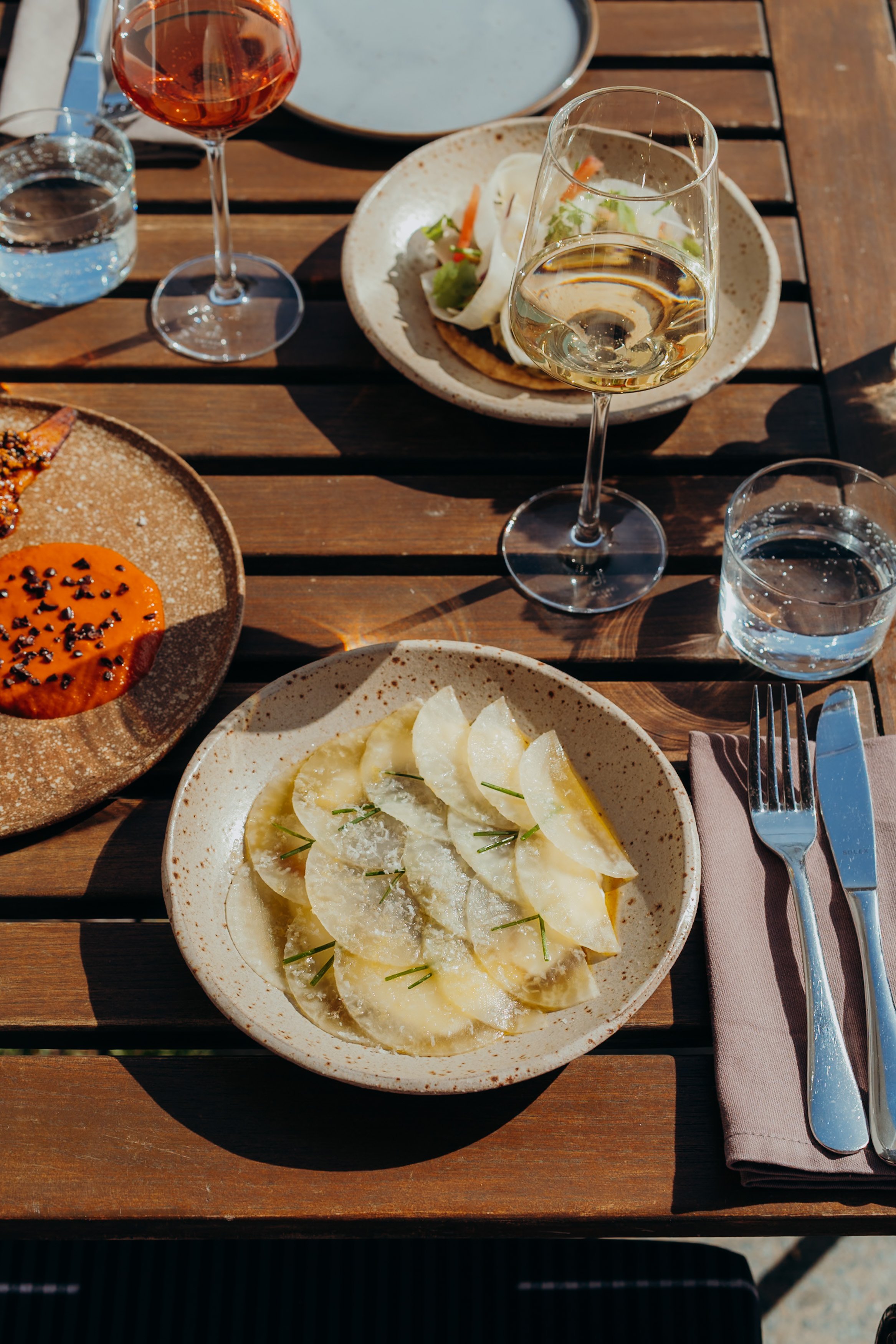 Ein Abendessen auf einem Holztisch mit mehreren Tellern und Gläsern, darunter ein Glas Weißwein und ein Glas Rosé. Im Mittelpunkt ein Teller mit dünnen, halbmondförmigen Scheiben eines weißen Gemüses, garniert mit Rosmarin, und ein Baquette. Weitere 