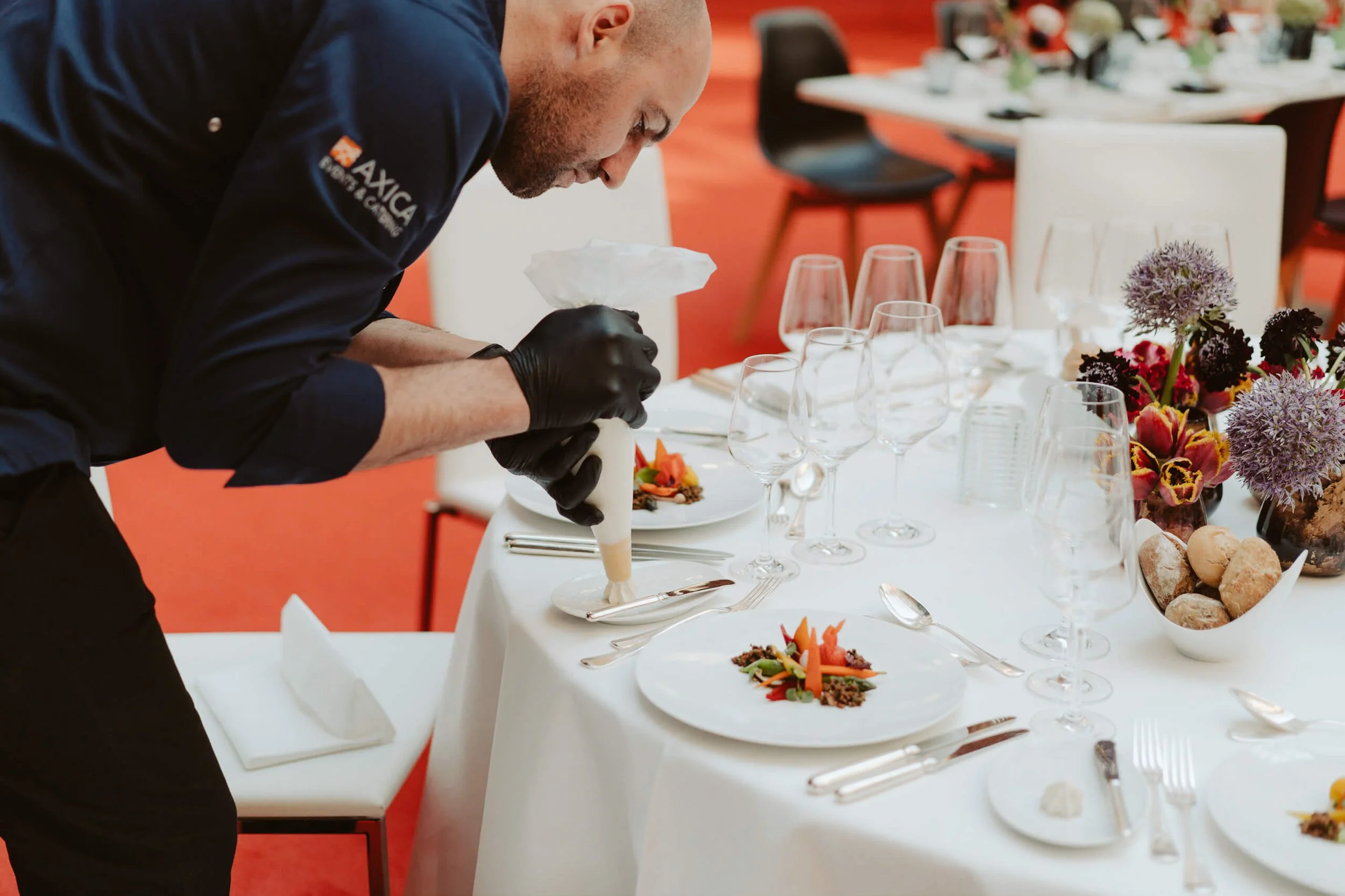 Ein Koch dekoriert ein festliches gedecktes Esstisch mit Gläsern, Blumen und verschiedenen Speisen bei einem Event.