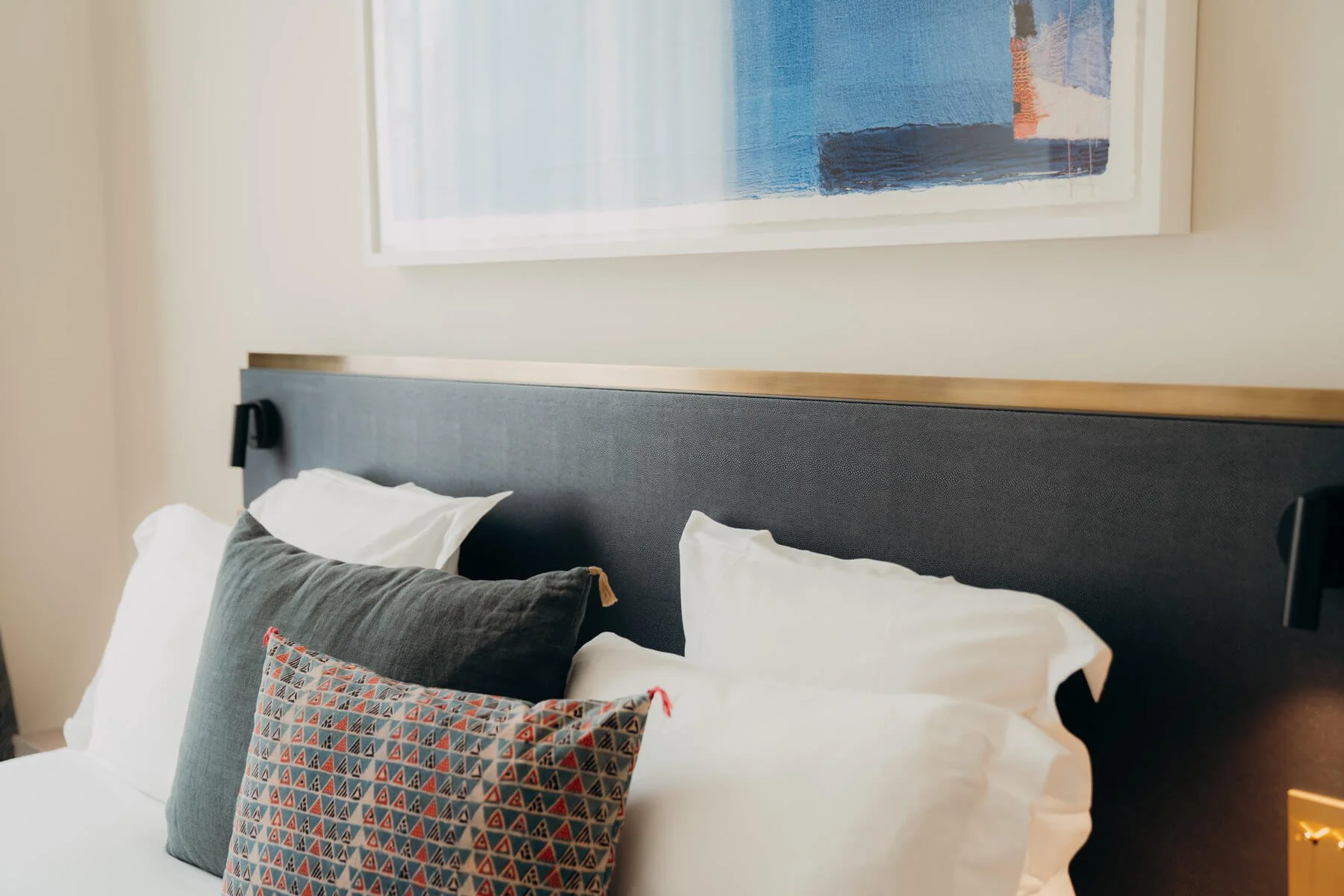 Bett mit weißen Bettlaken und mehreren Kissen, darunter ein graues Kissen und ein Kissen mit geometrischem Muster, vorm Fenster im Schlafzimmer.