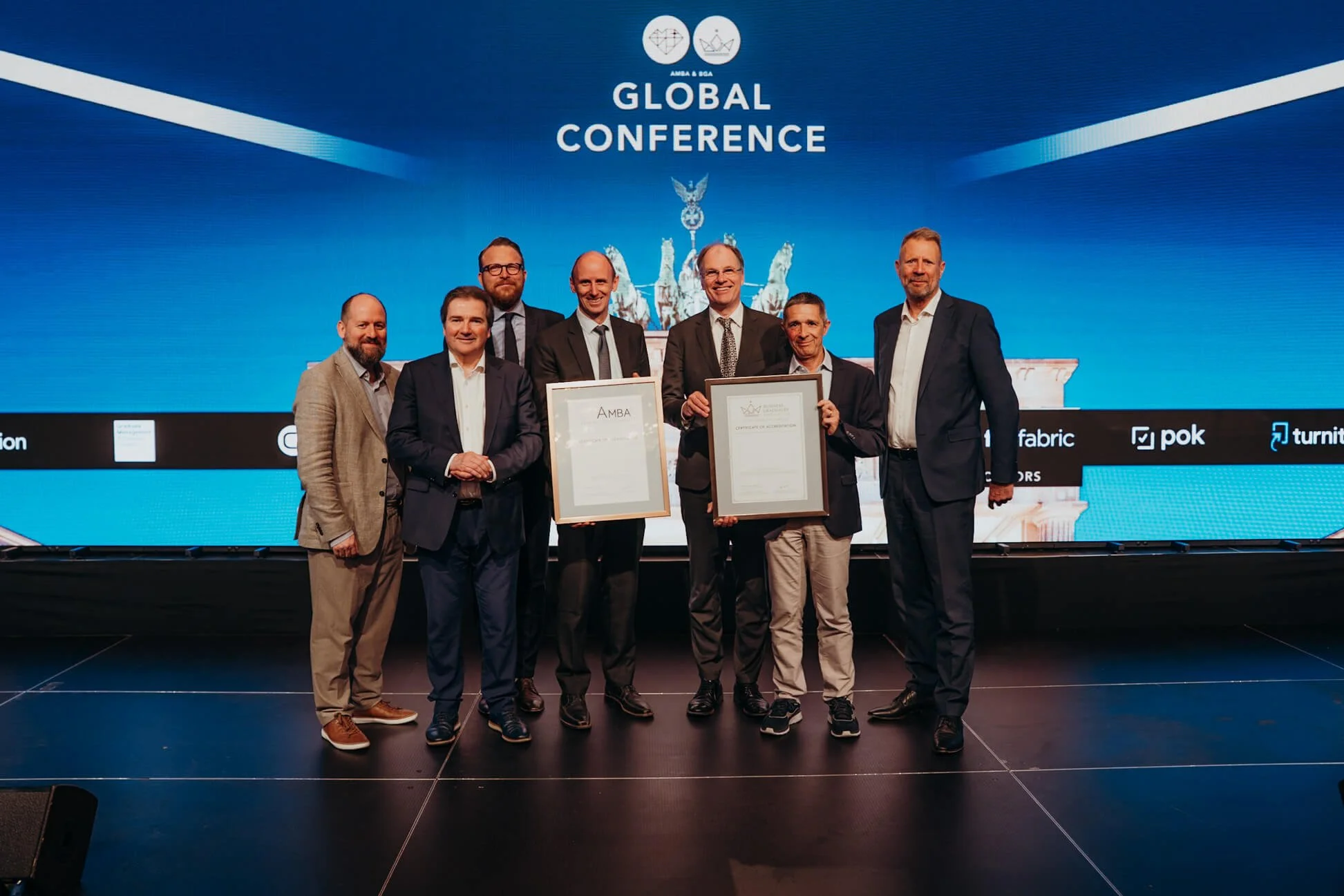 Group of seven men standing on stage at a conference, holding framed certificates, with a large blue screen behind them displaying "Global Conference" and the U.S. Capitol statue.