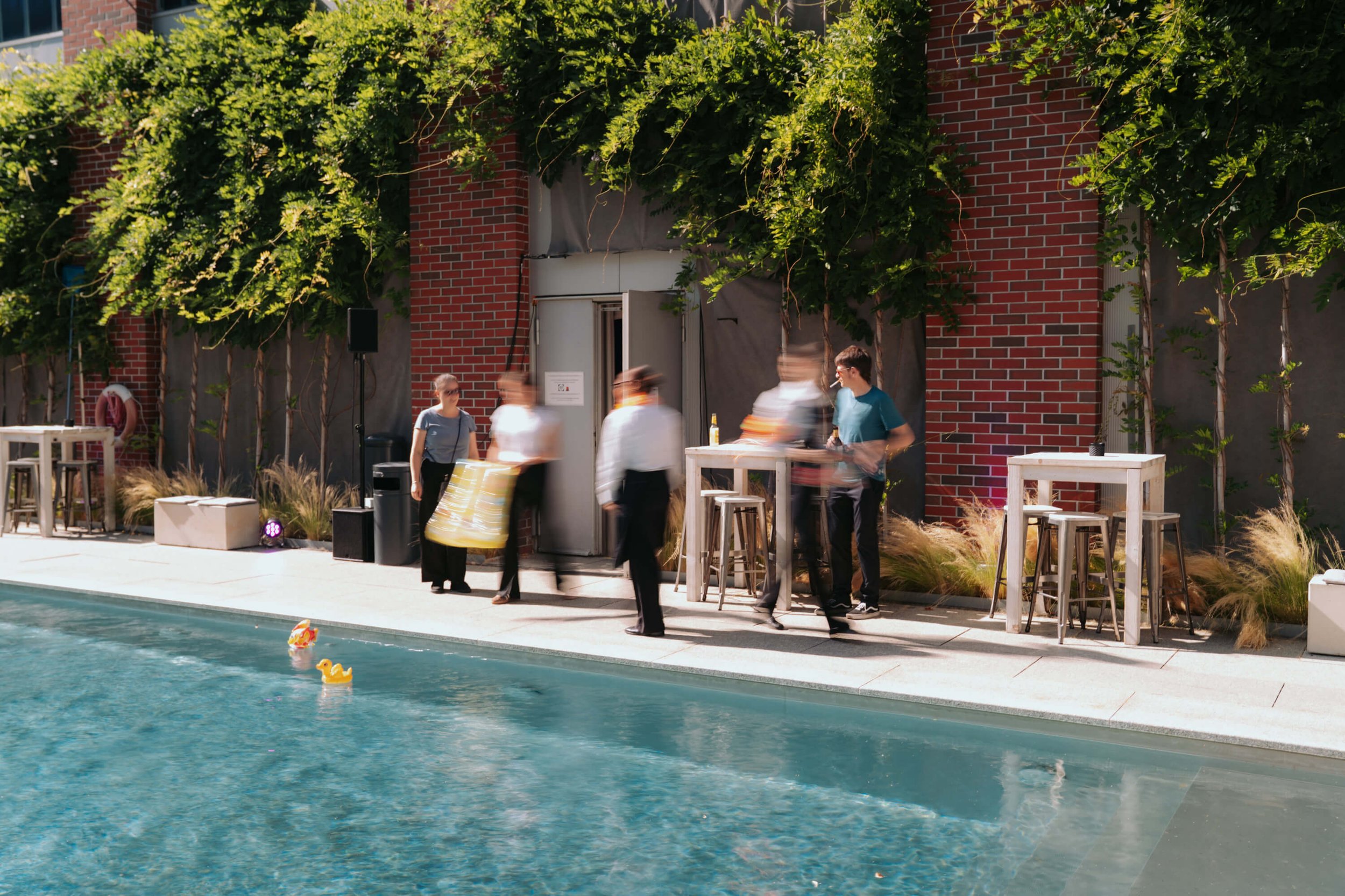 Menschen am Pool bei einer Poolparty, einige stehen an Stehtischen, andere sind im Wasser, im Hintergrund eine Hauswand mit bewachsenen Fenstern, sonniges Wetter.