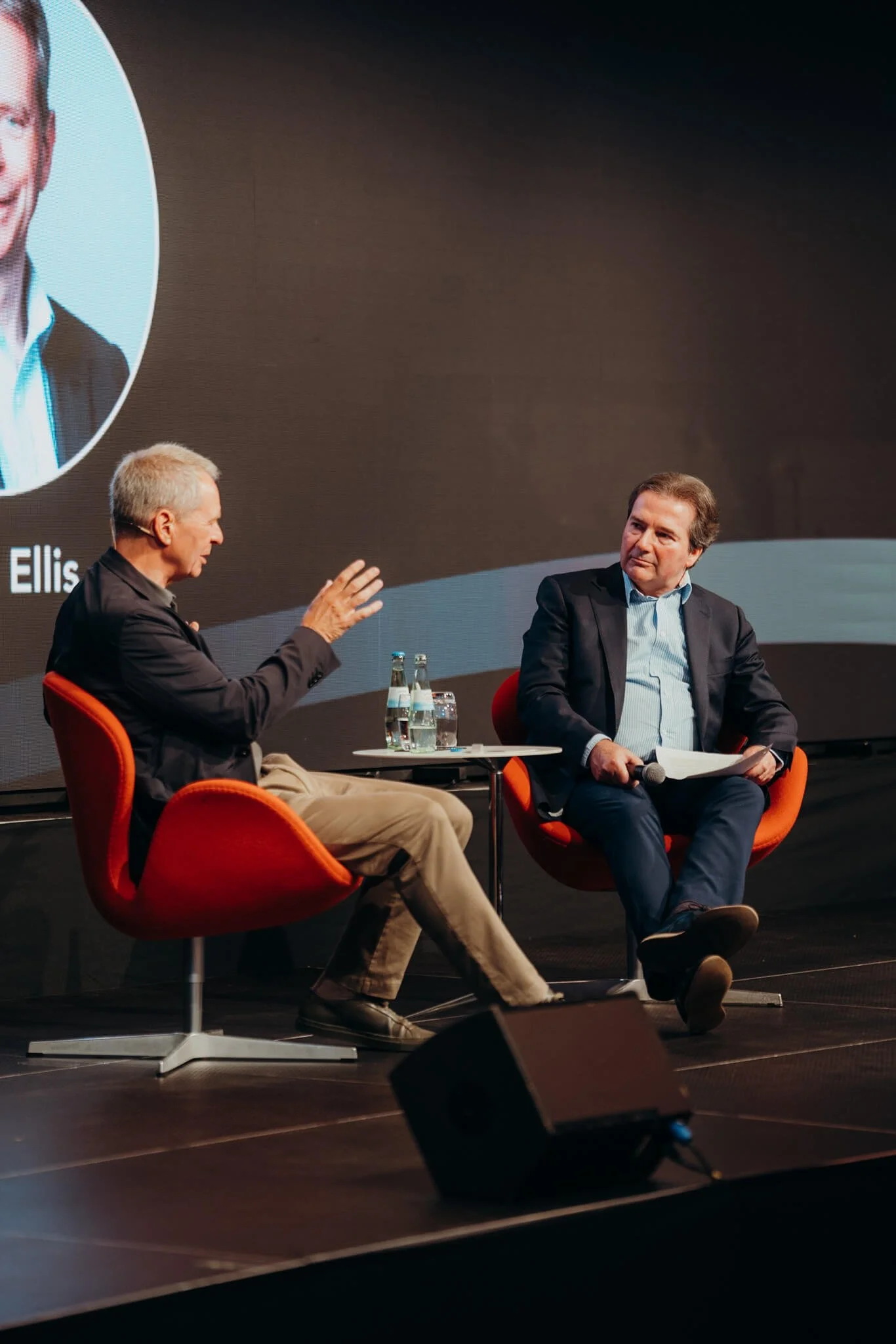 Two men sitting on orange chairs, engaged in a conversation on stage during a panel discussion or interview. There are three bottles of water on a small table between them. One man is gesturing with his hand while speaking, and the other is listening