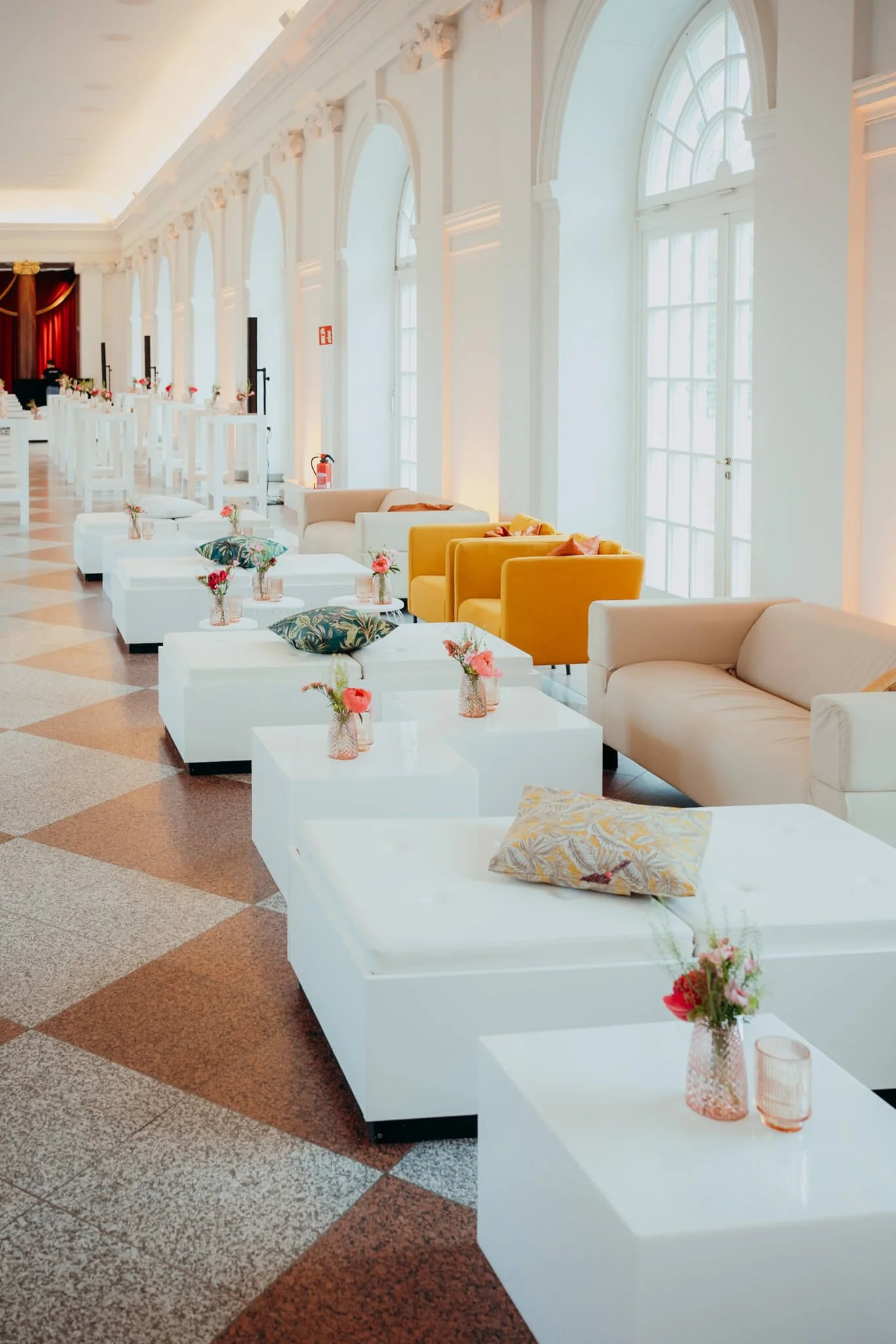Bright, elegant lounge area with large arched windows, white sofas, yellow armchairs, white coffee tables with pink flowers, decorative pillows, and a checkered tile floor.