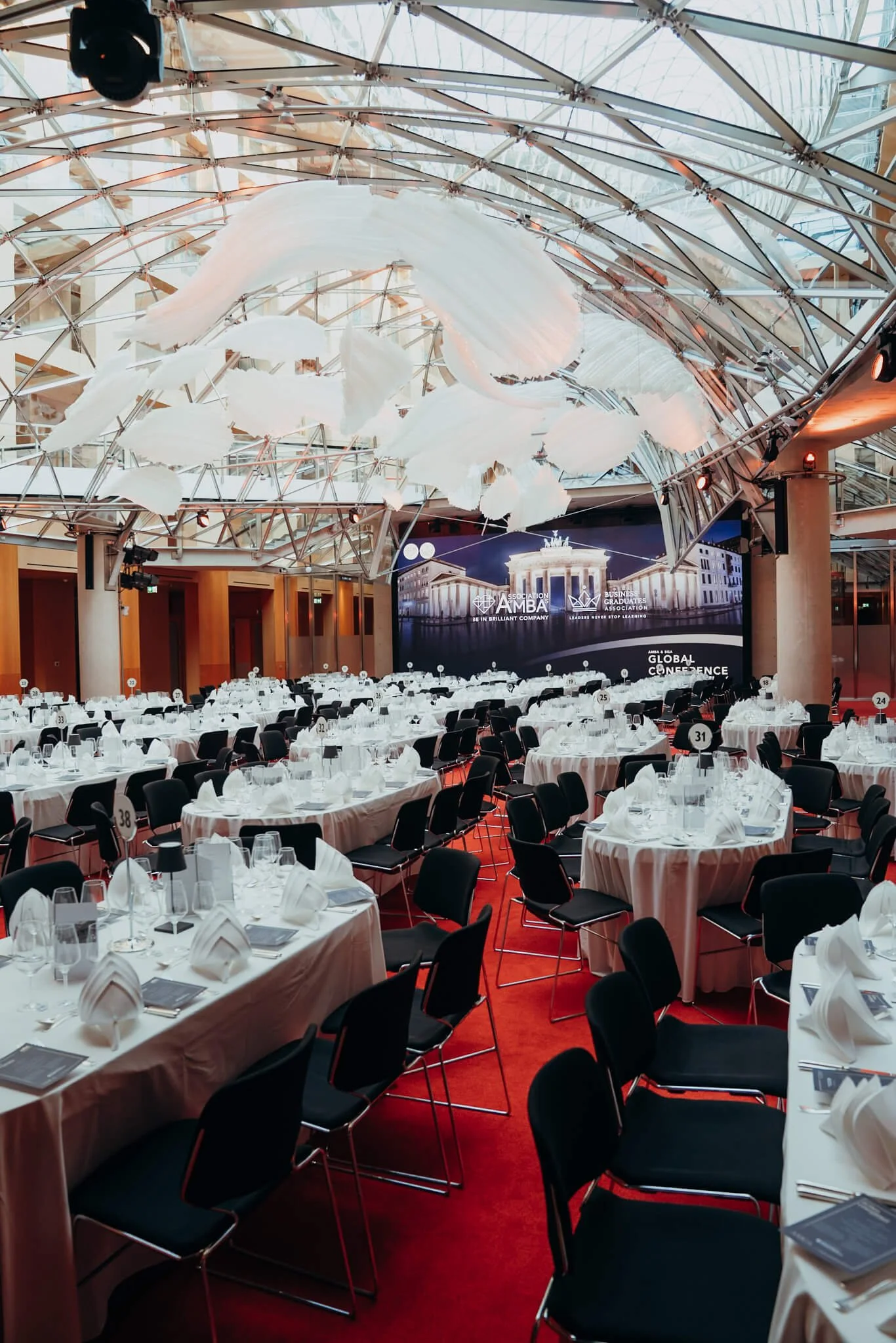 A large banquet hall with round and rectangular tables set with white tablecloths, neatly folded napkins, glasses, and silverware, all arranged for a formal event. The hall has a modern, glass arched ceiling and a big digital screen at the front disp