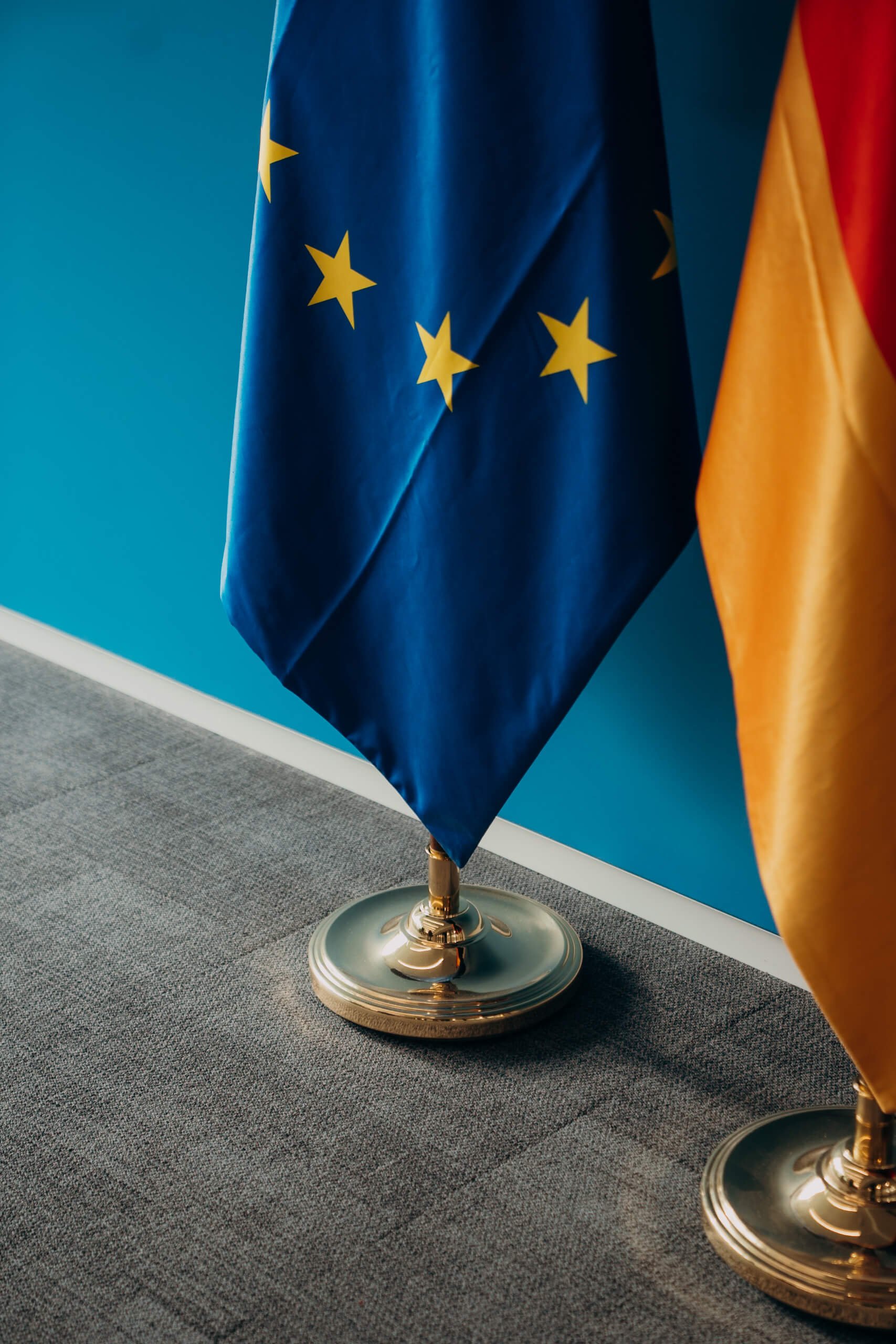 Nahaufnahme von zwei Flaggen, einer mit den europäischen Sternen auf blauem Hintergrund und einer mit den deutschen Farben, auf einem Messestand, mit Teppichboden darunter.