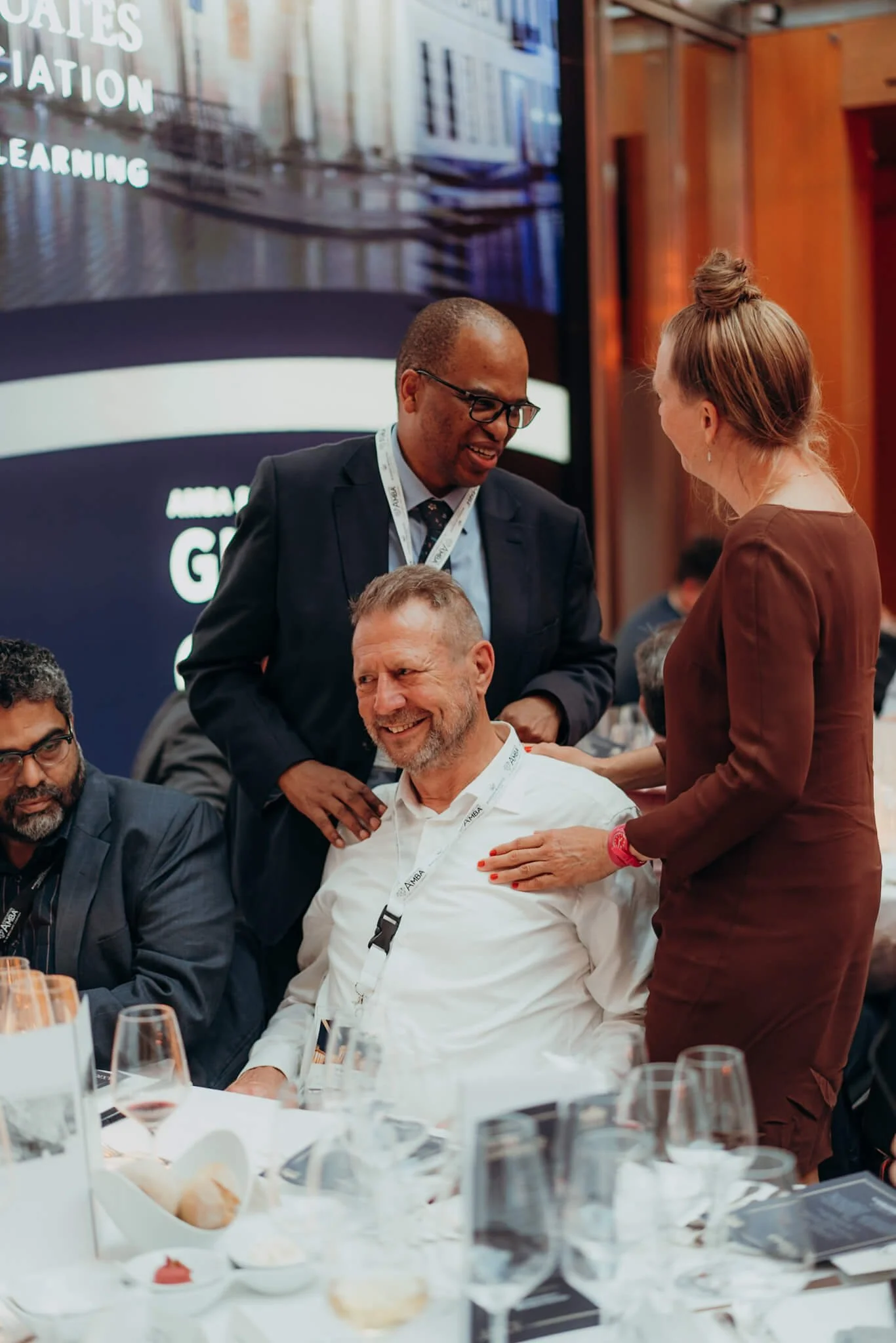 People engaged in a conversation at a formal event, with a man in a suit and glasses smiling and a woman with a bun talking to him, while others sit at the table.