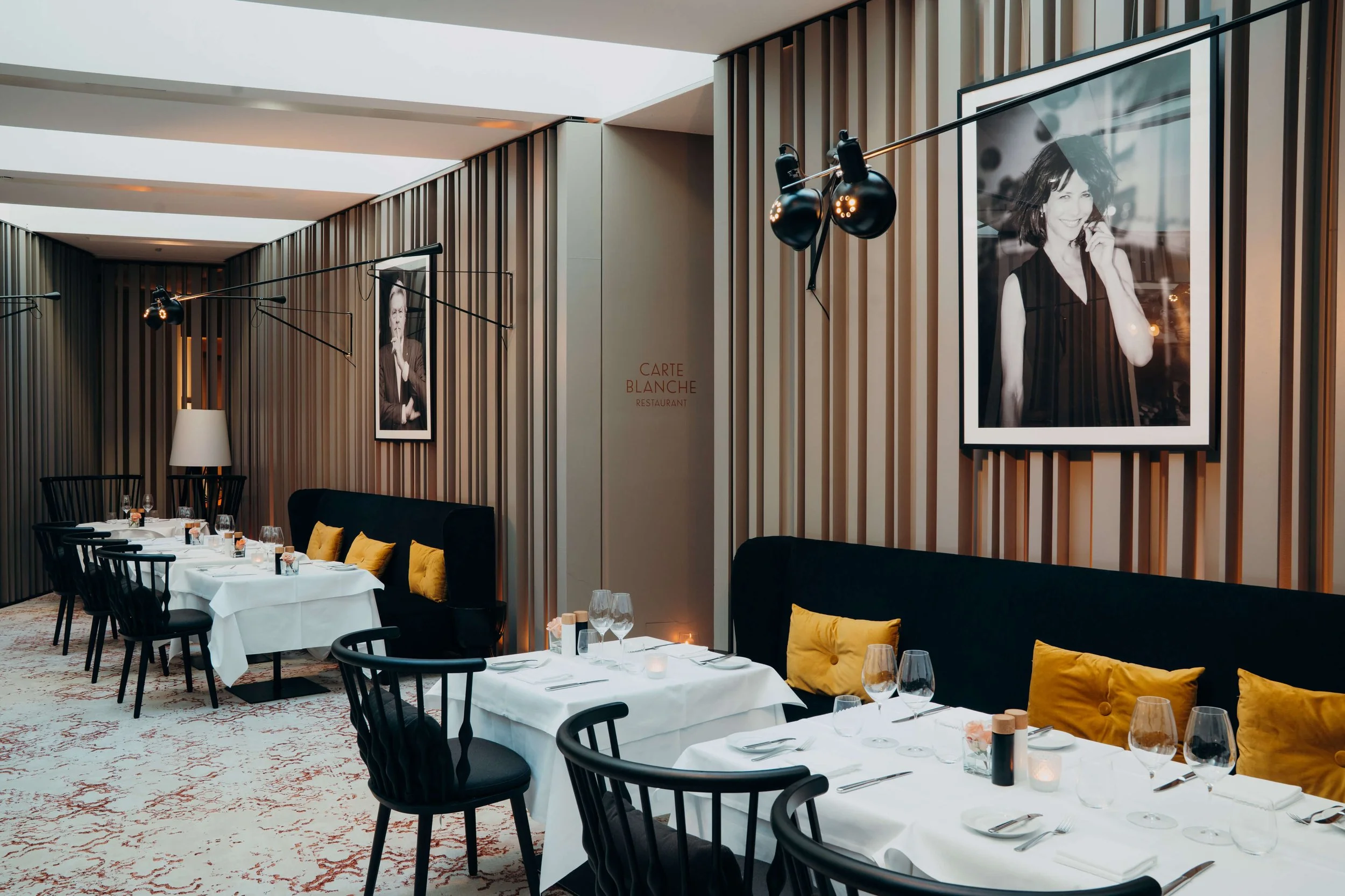 Elegant restaurant dining room with white tablecloths, black chairs, and yellow pillows on black banquettes. Framed black-and-white photographs on the wood-paneled walls, modern lighting fixtures, and a sign that reads 'Carte Blanche Restaurant'.