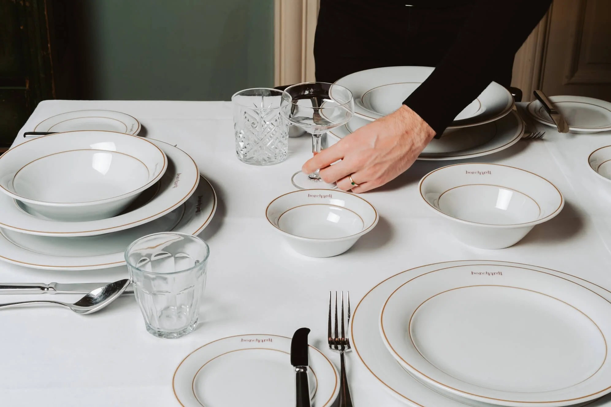 A person arranging clear glassware and white dinnerware with gold trim on a white tablecloth, with individual plates, bowls, knives, forks, spoons, and glasses.