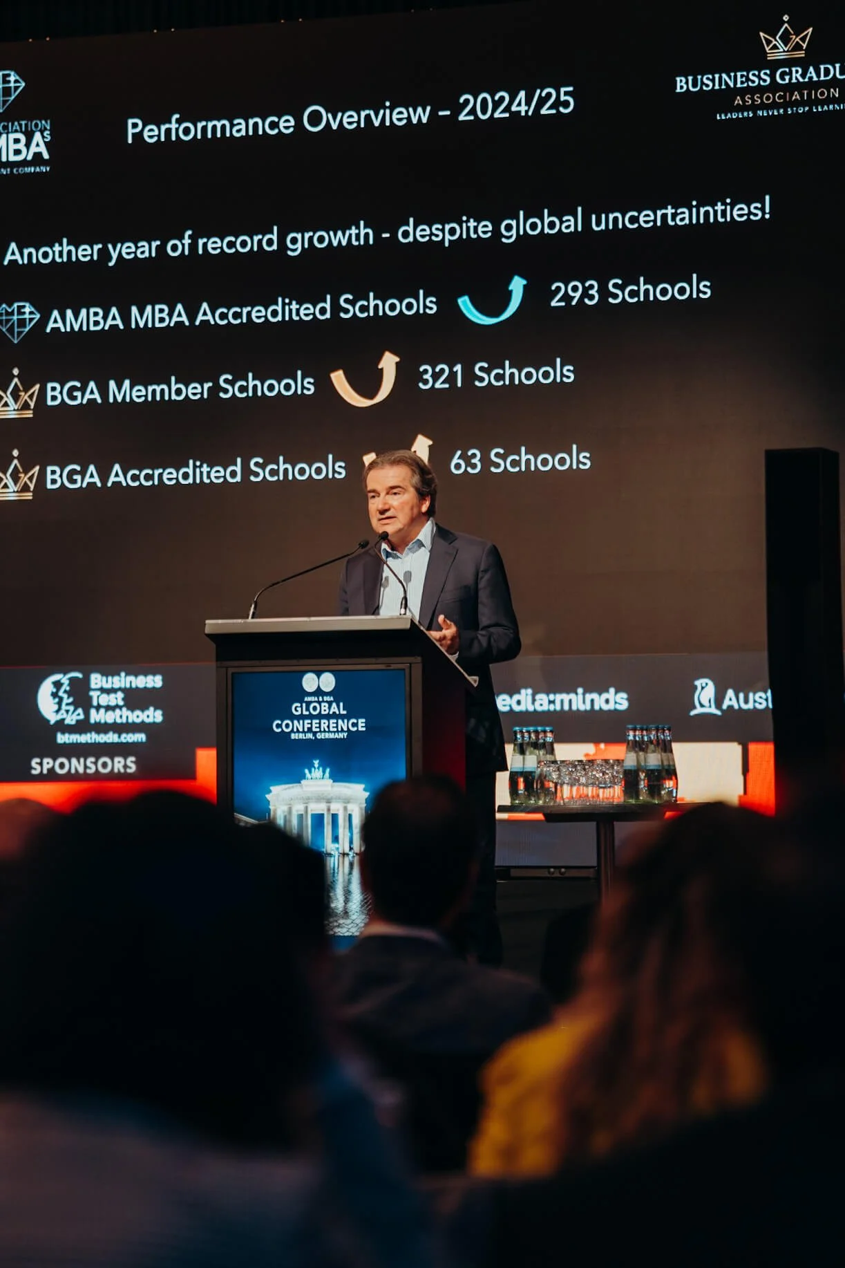A man in a suit giving a presentation at a conference, with a slide displaying performance statistics for 2024/25 behind him. The slide mentions record growth, 293 AMBA MBA accredited schools, 321 BGA member schools, and 63 BGA accredited schools.