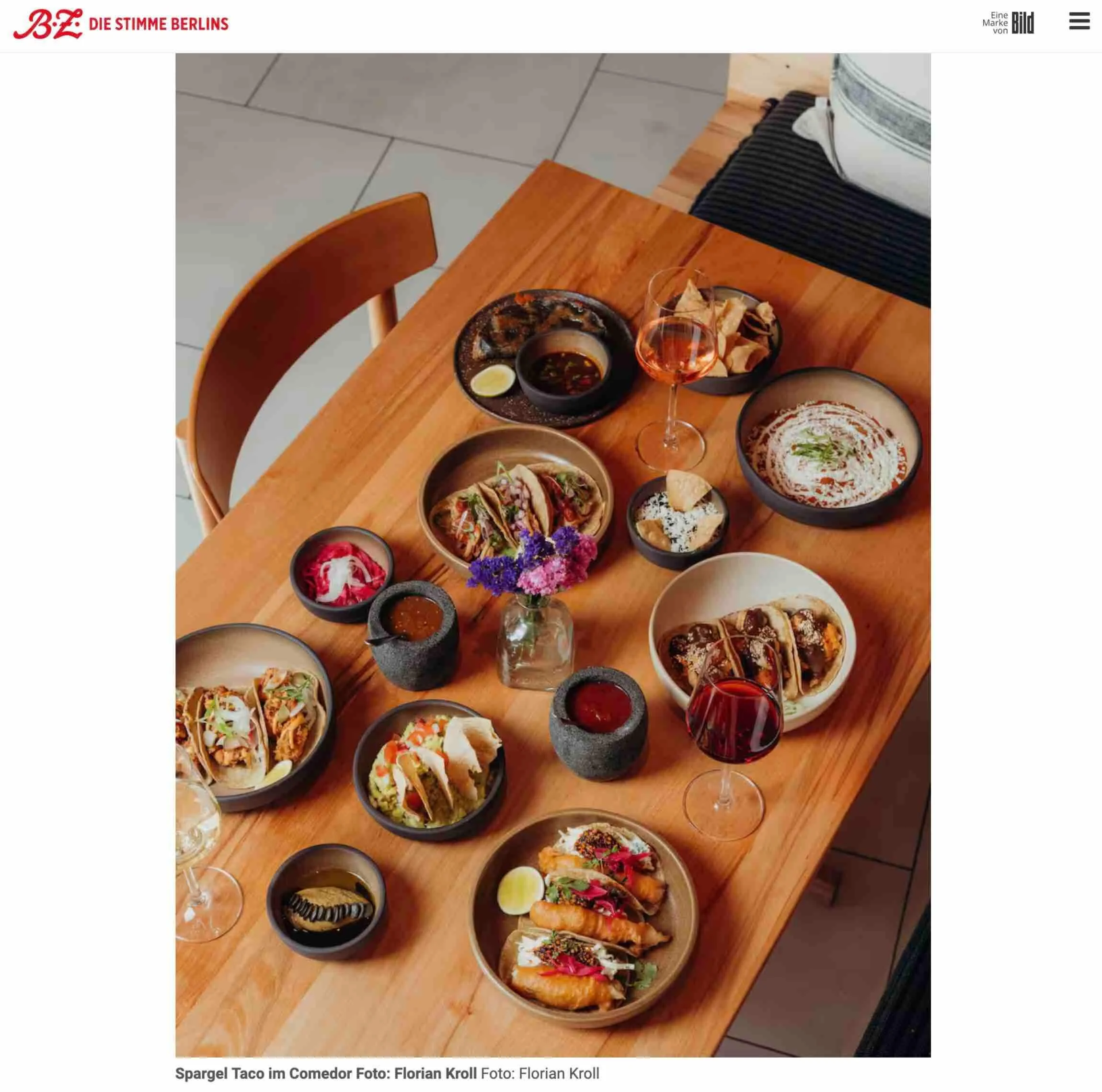 Ein Holztisch mit mehreren Schalen und Gläsern, die verschiedene Speisen und Getränke enthalten. Es gibt Tacos, ein Glas Wein, und kleine Schälchen mit Soßen und Beilagen. Im Hintergrund sind weitere Tacos und Dekorationen sichtbar.