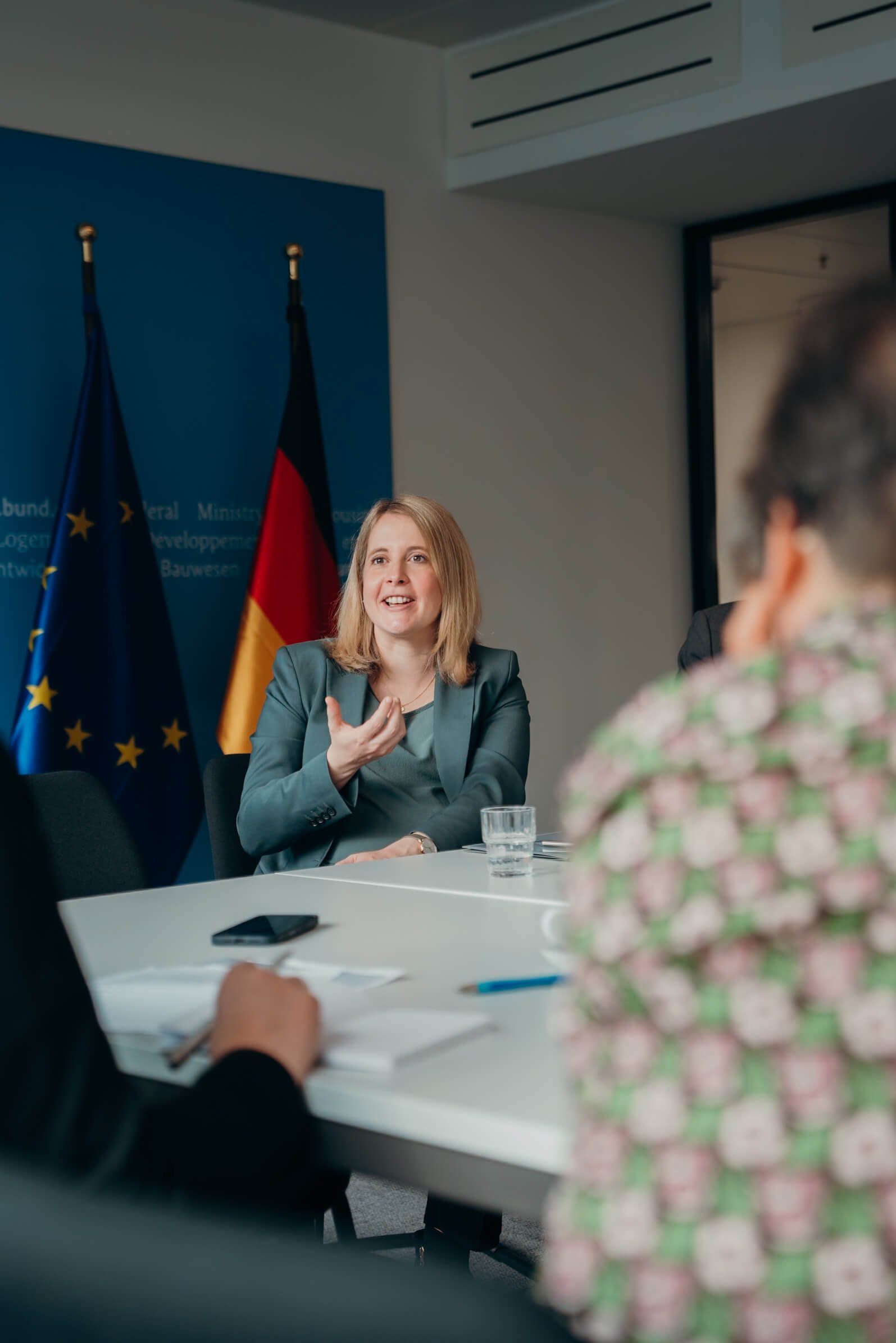 Eine Frau in einem grauen Blazer spricht während einer Besprechung in einem Konferenzraum, im Hintergrund die deutschen und europäischen Flaggen.