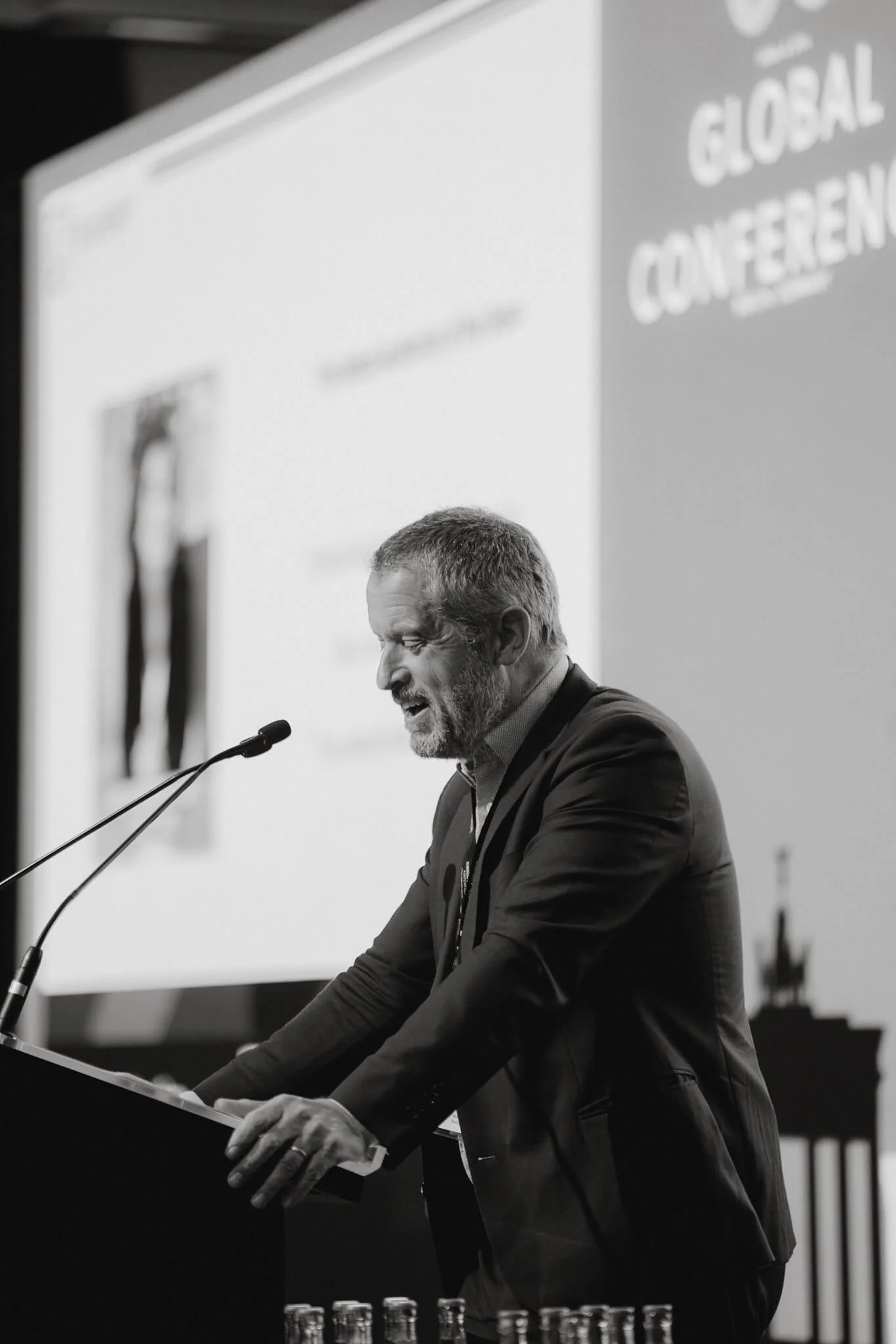 A man with gray hair and beard giving a speech at a conference, standing at a podium with a microphone, in front of a large presentation screen.