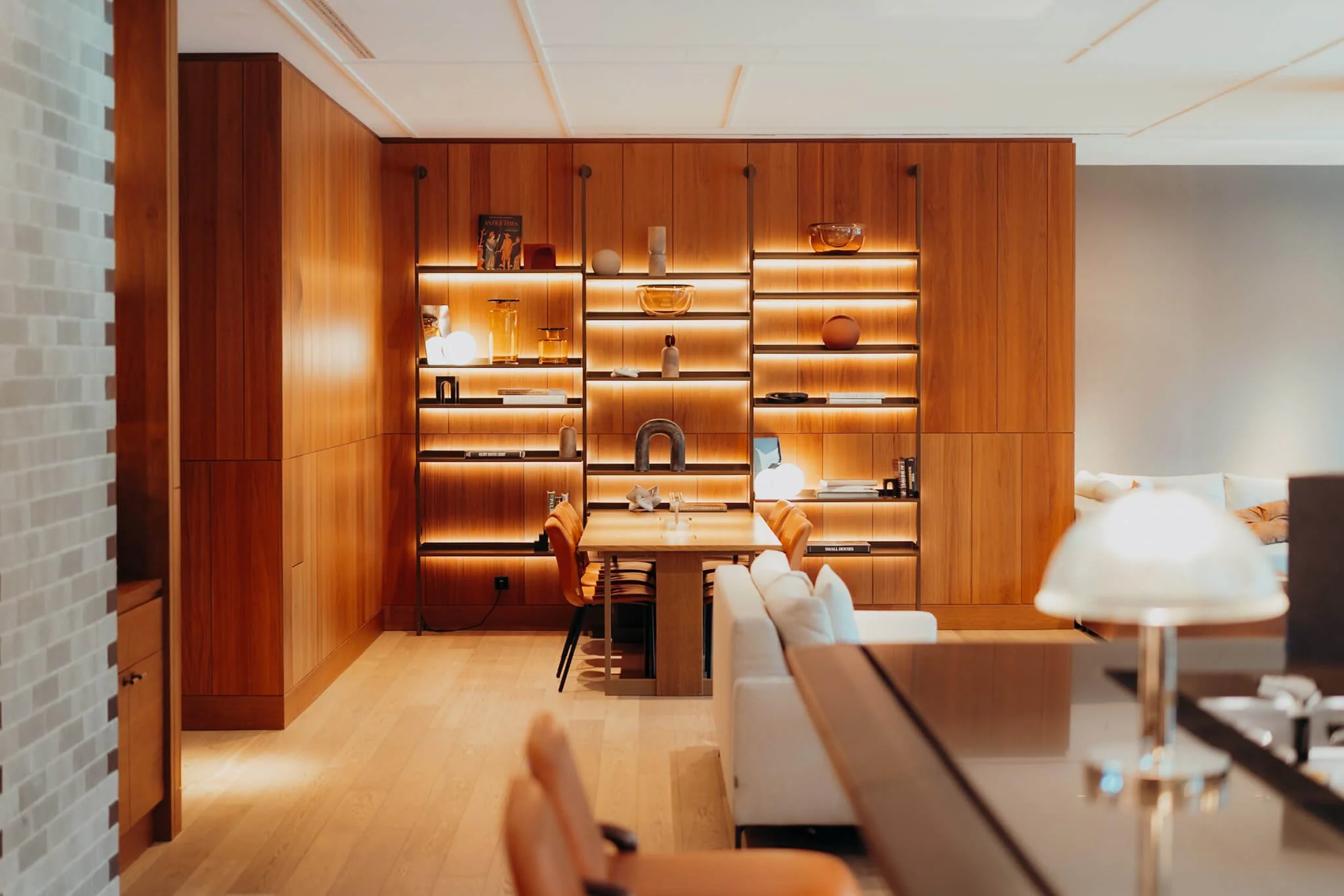 A cozy, well-lit living room with a wooden wall unit filled with various decorative objects. A small dining table with orange chairs is in front of the wall unit, and a white sofa is partially visible on the right side of the image.