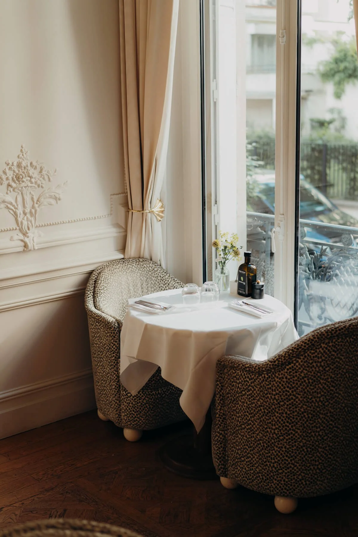 Ein gemütlicher Speiseraum mit einem runden Tisch, gedeckt mit einer weißen Tischdecke, Besteck und Gläsern. Zwei gepolsterte Armlehnstühle mit leopardähnlichem Muster stehen an den Tischen. Das Fenster im Hintergrund lässt Tageslicht herein, und auf