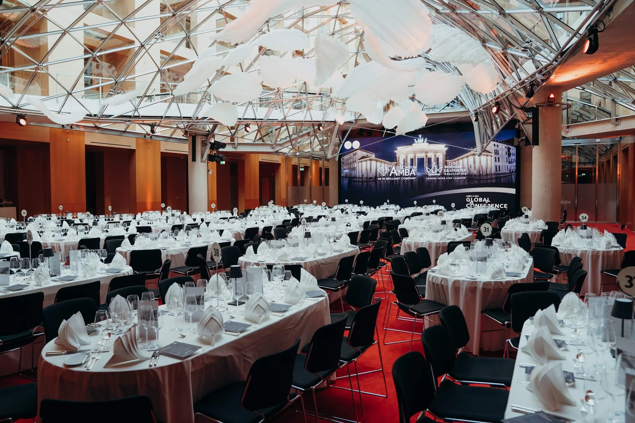 A large event hall set up with round tables covered in white tablecloths, elegant glassware, and folded white napkins, ready for a formal conference or banquet, with a large screen displaying event details at the front.