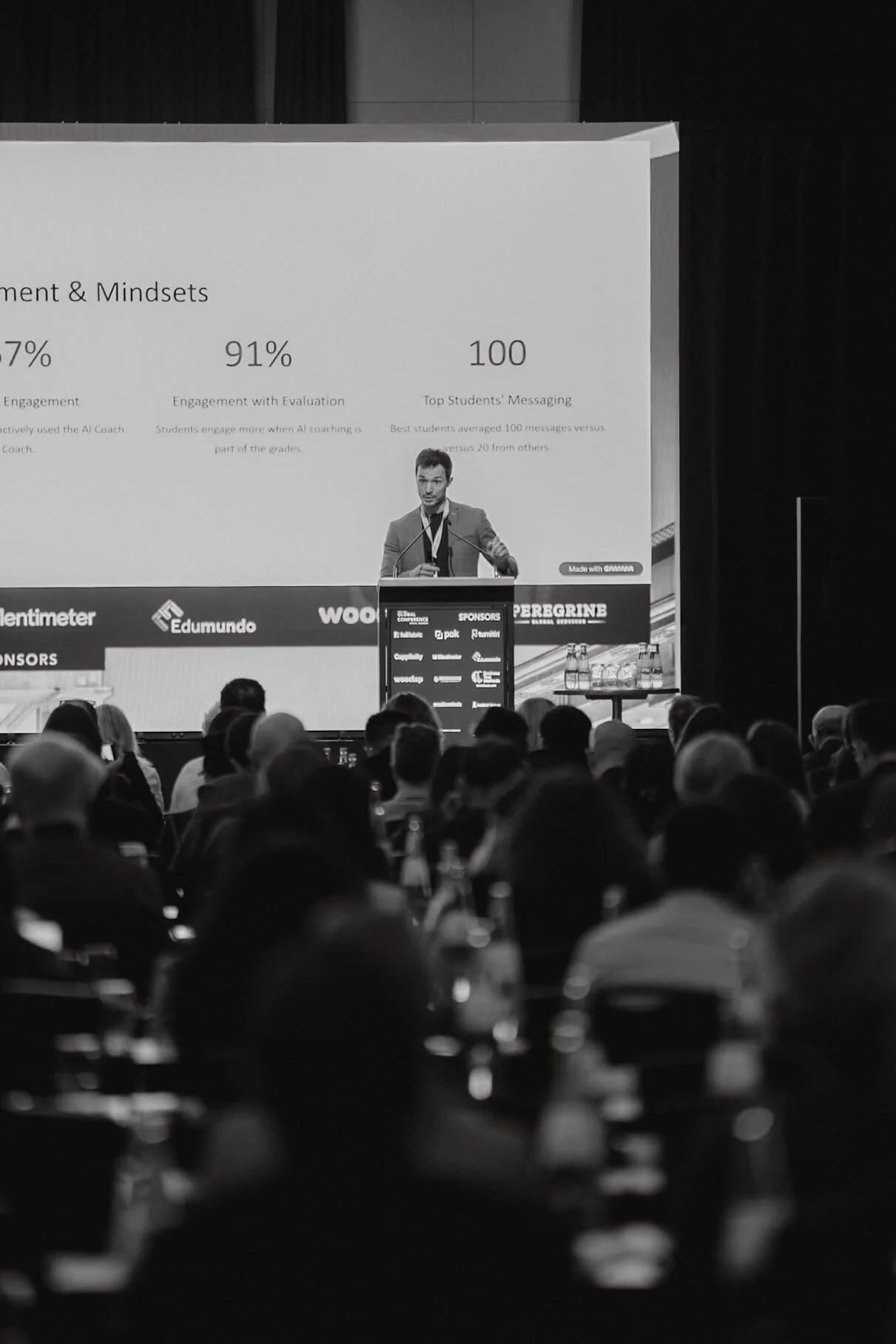 A black-and-white photo of a speaker at a conference, standing behind a podium on a large stage with a projection screen behind him. The audience is seated and listening, with some water bottles visible on the tables.