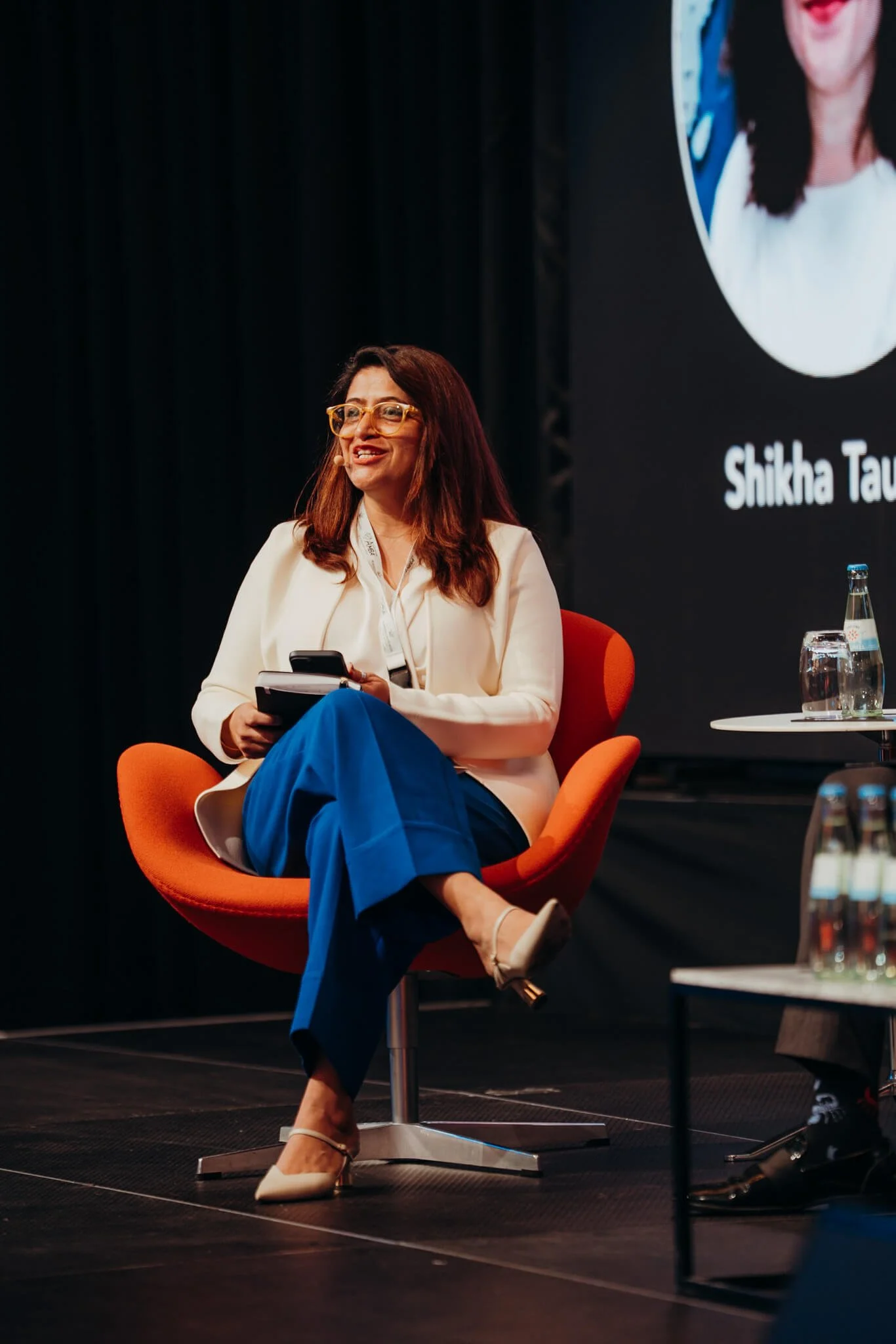 A woman with brown hair and glasses sitting on an orange chair on stage, holding a notebook and a phone, wearing a white blazer, blue pants, and beige heels, smiling during a panel discussion.