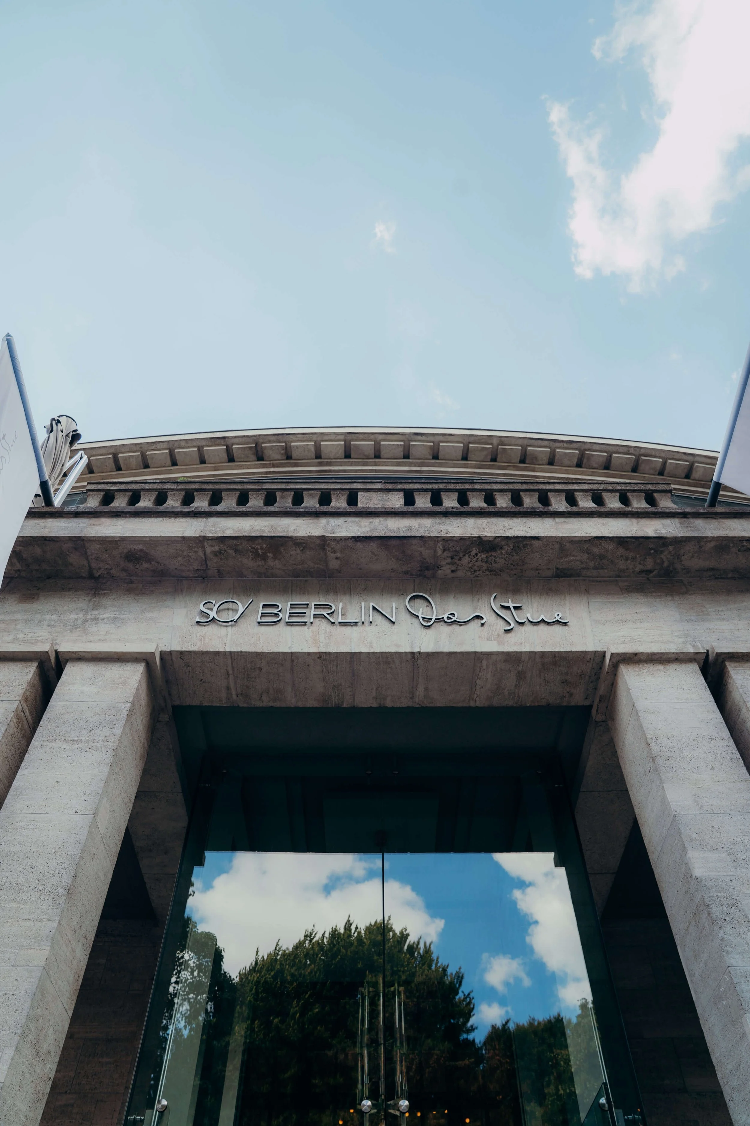 The entrance to a building with the sign reading 'SO BERLIN Das Stue' above the glass door, with reflections of trees and blue sky with clouds.