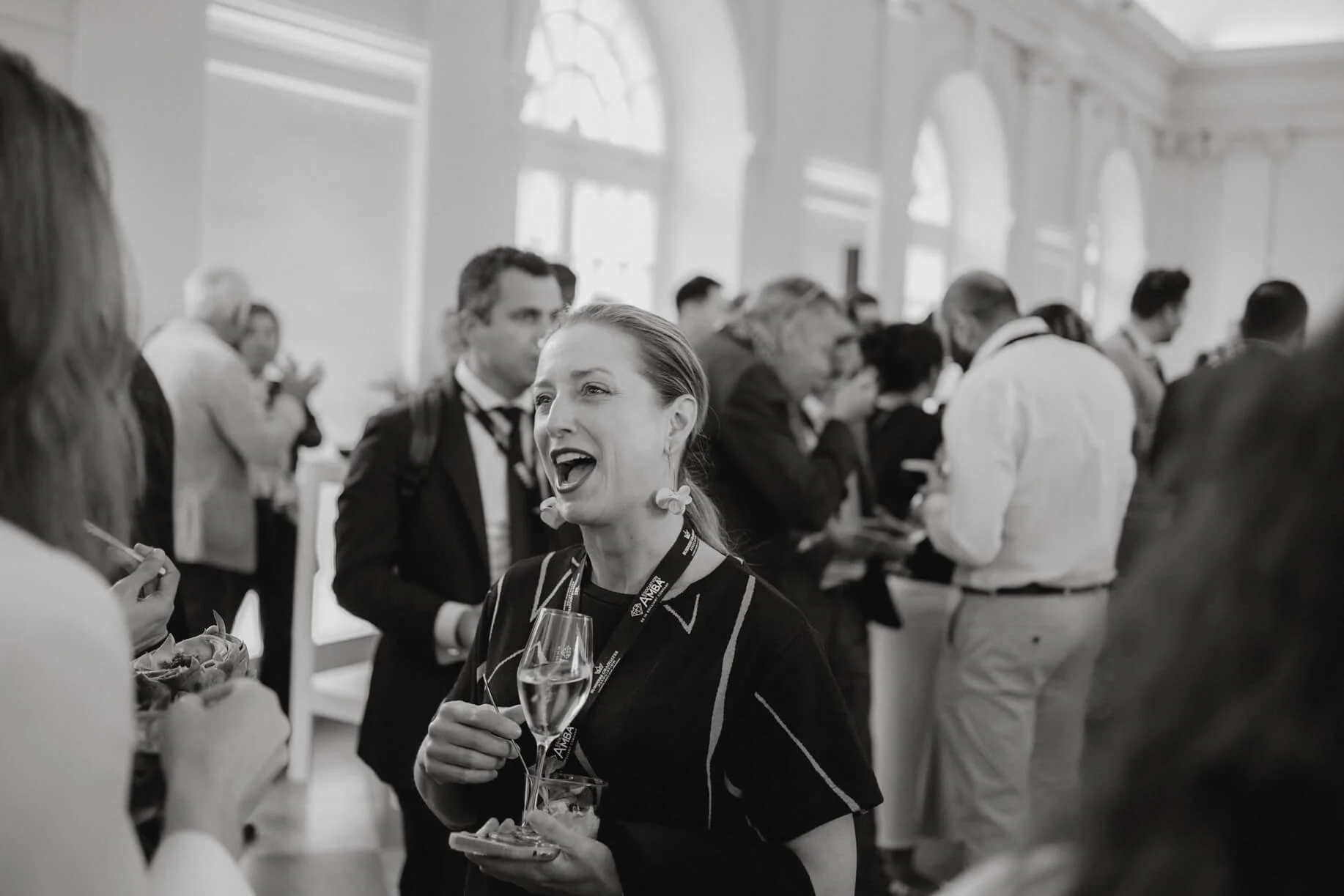 A woman in a black shirt is holding a glass of champagne and talking to another person at a professional event or conference. The background shows a professional gathering with people engaging in conversations in a large, elegant room with high windo