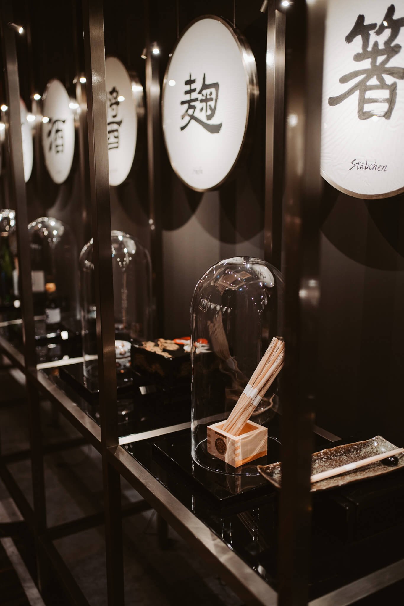 A display shelf with glass coverings in a Japanese restaurant, showcasing chopsticks in a wooden holder, black trays, and sushi dishes. Decorative panels with Japanese characters are above the shelf.