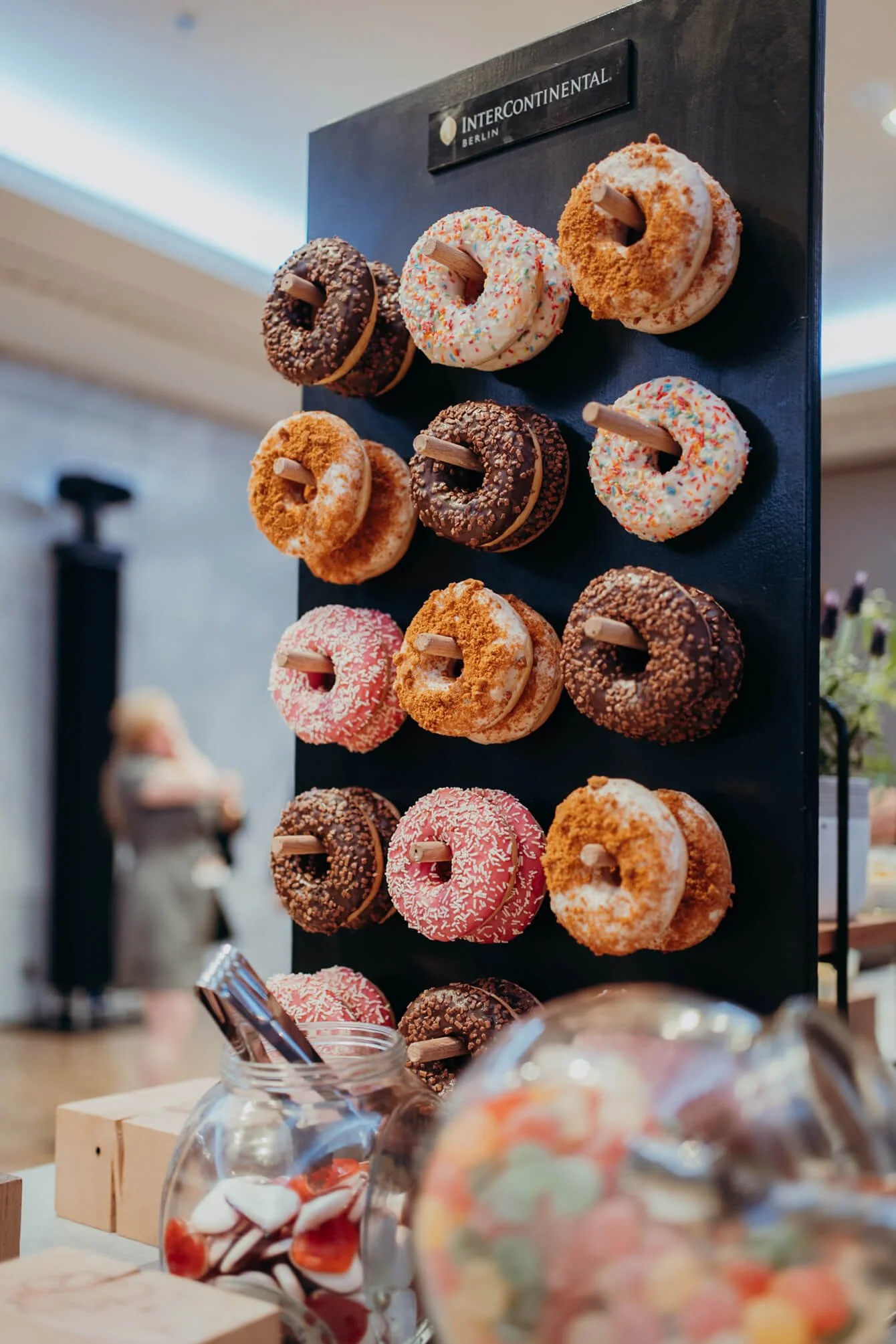 Display of assorted donuts on a black stand labeled 'Intercontinental Berlin,' with various toppings like sprinkles and chocolate, and jars of candies in the foreground.