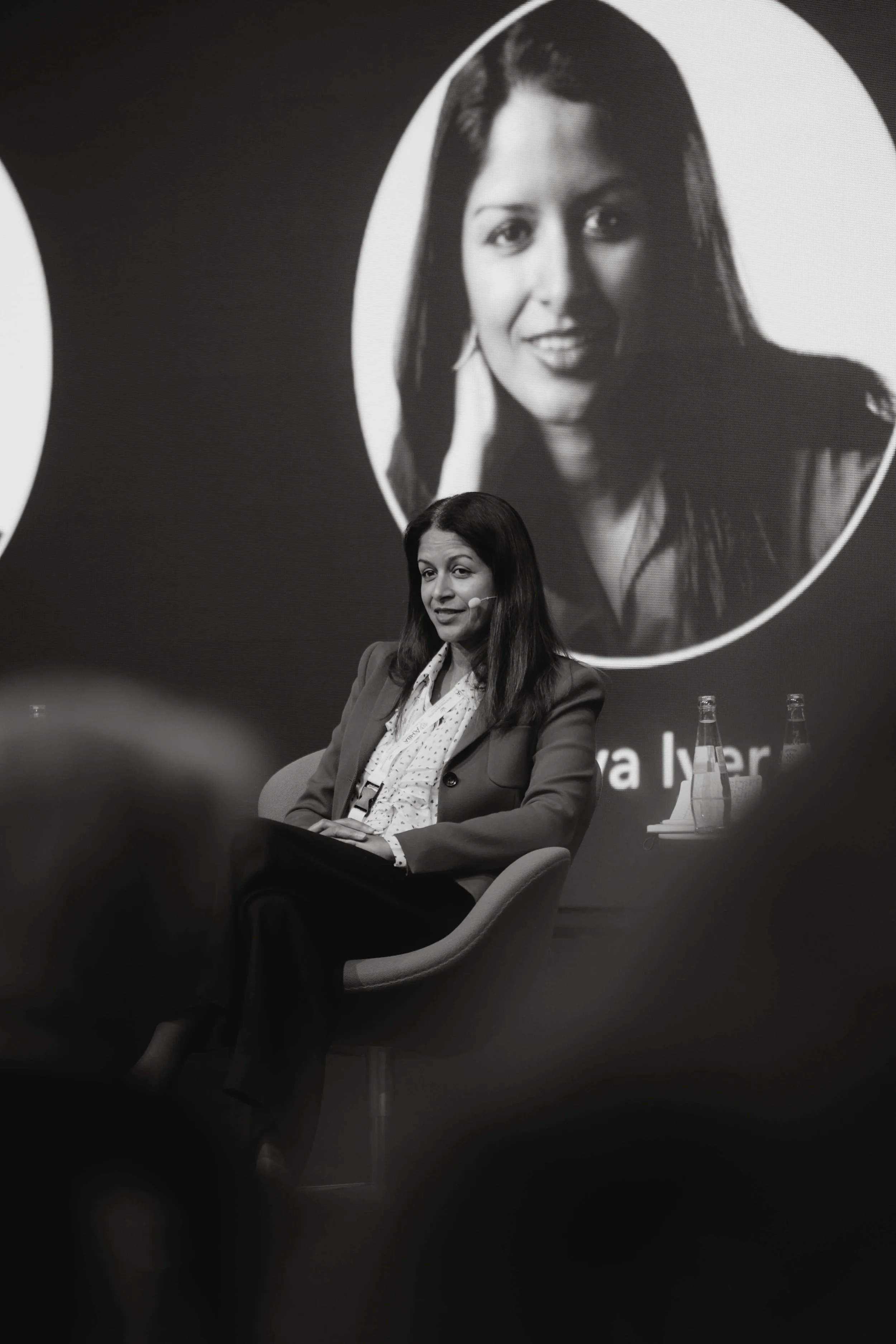 Black and white photo of a woman speaking at a panel, sitting in a chair with a microphone headset, in front of a screen displaying her large portrait.