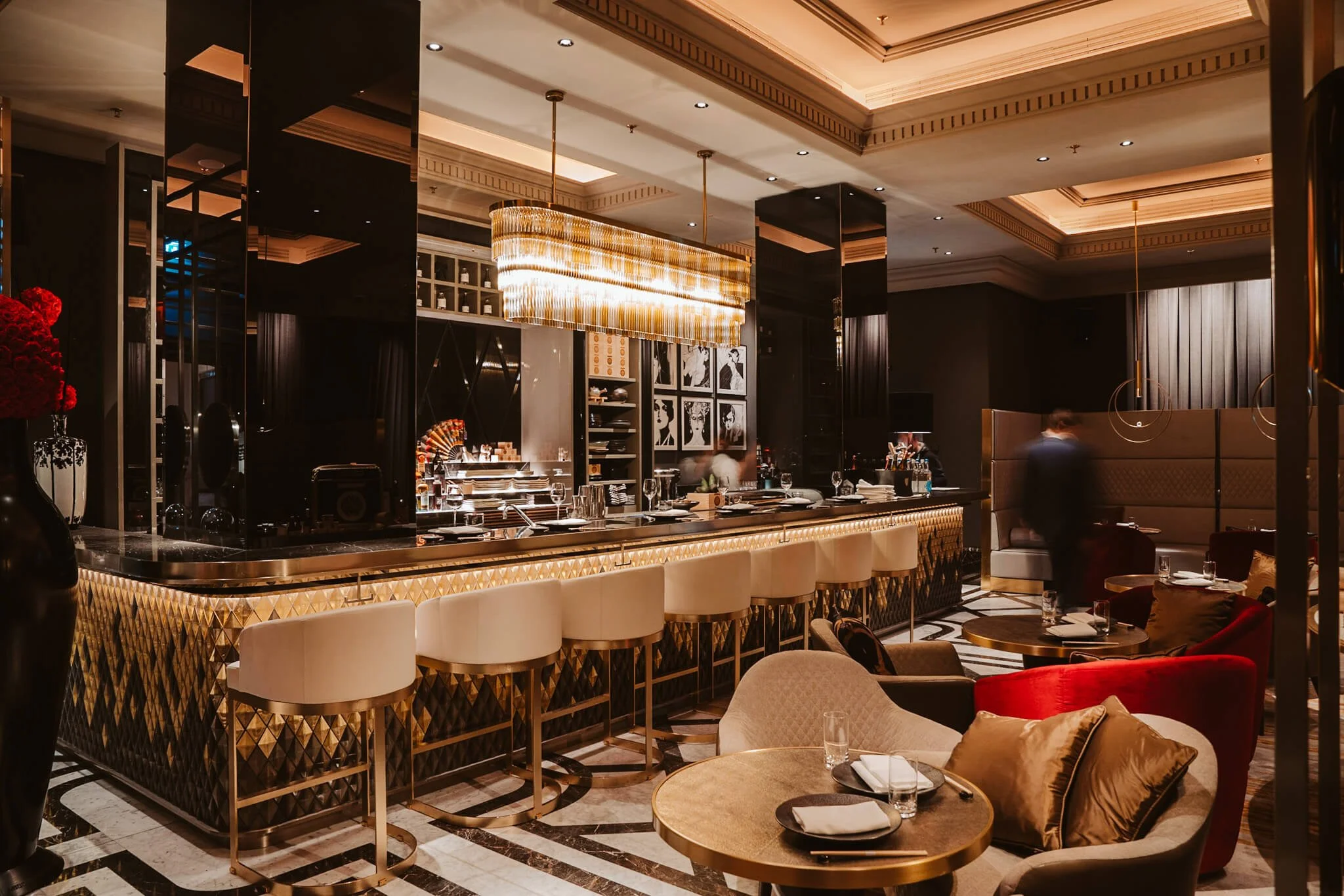 Interior of an upscale restaurant or bar with a marble countertop, cream-colored stools, and a gold chandelier. There's a seating area with plush chairs and a table set with glasses and napkins.