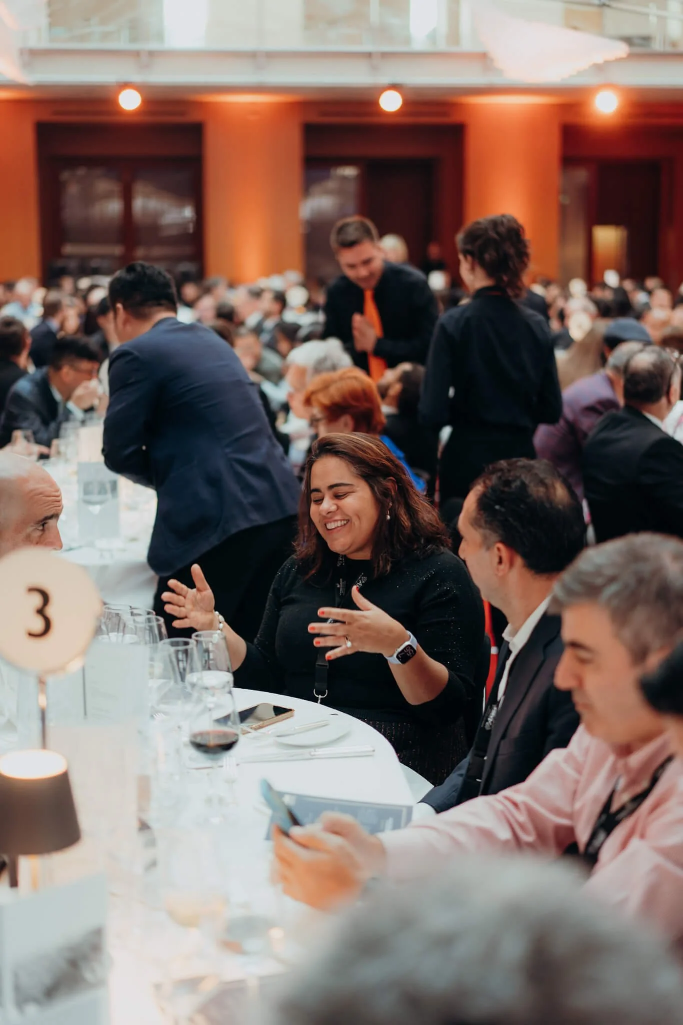A woman at a formal event, smiling and engaging in conversation with two men seated at a round table.