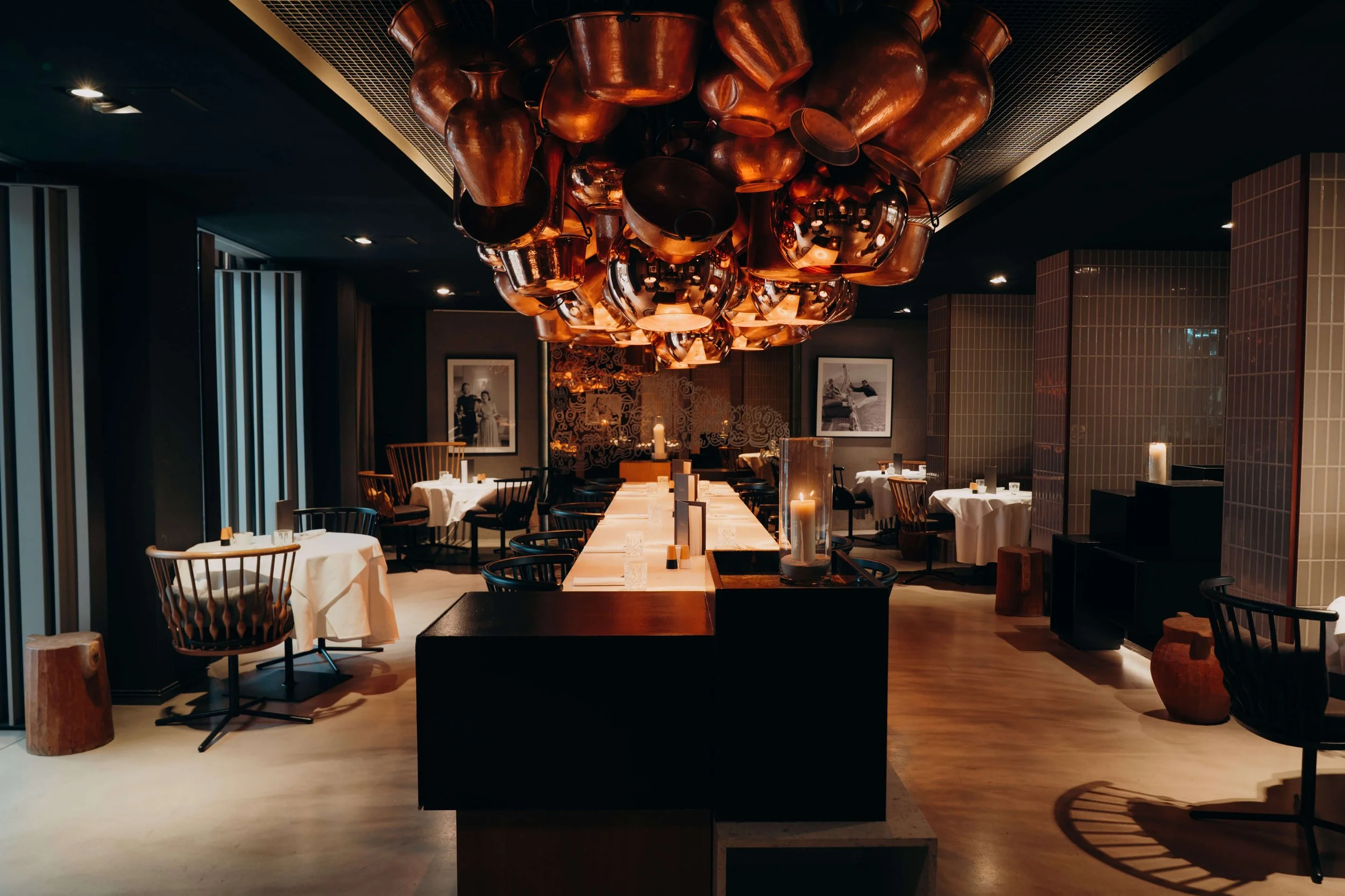 Interior of a modern restaurant with a dark ceiling, copper pendant light fixture, round tables with white tablecloths, candles, and black chairs with wooden armrests.