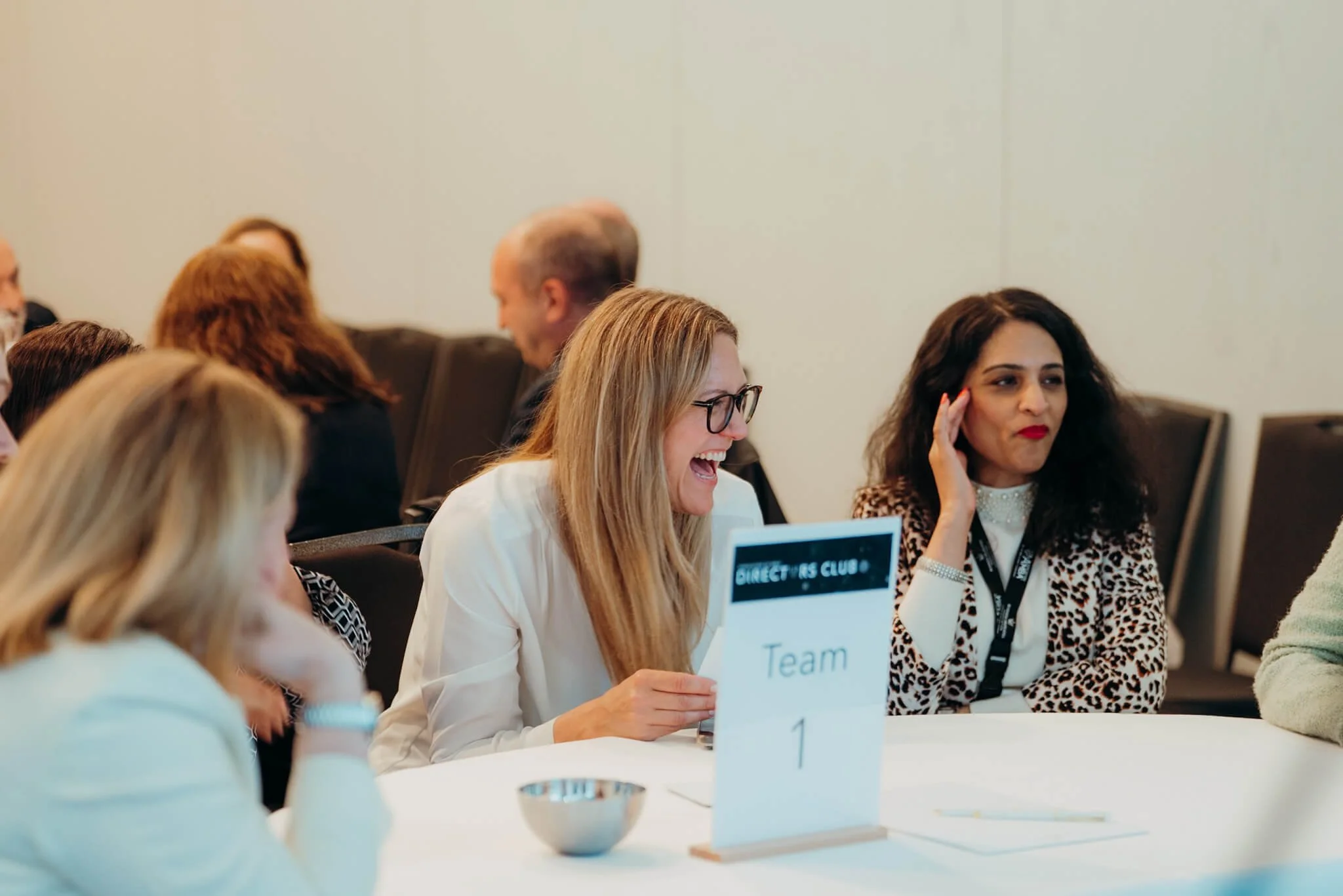 Group of people at a conference table, with a sign reading 'Team 1', smiling and engaging in conversation.