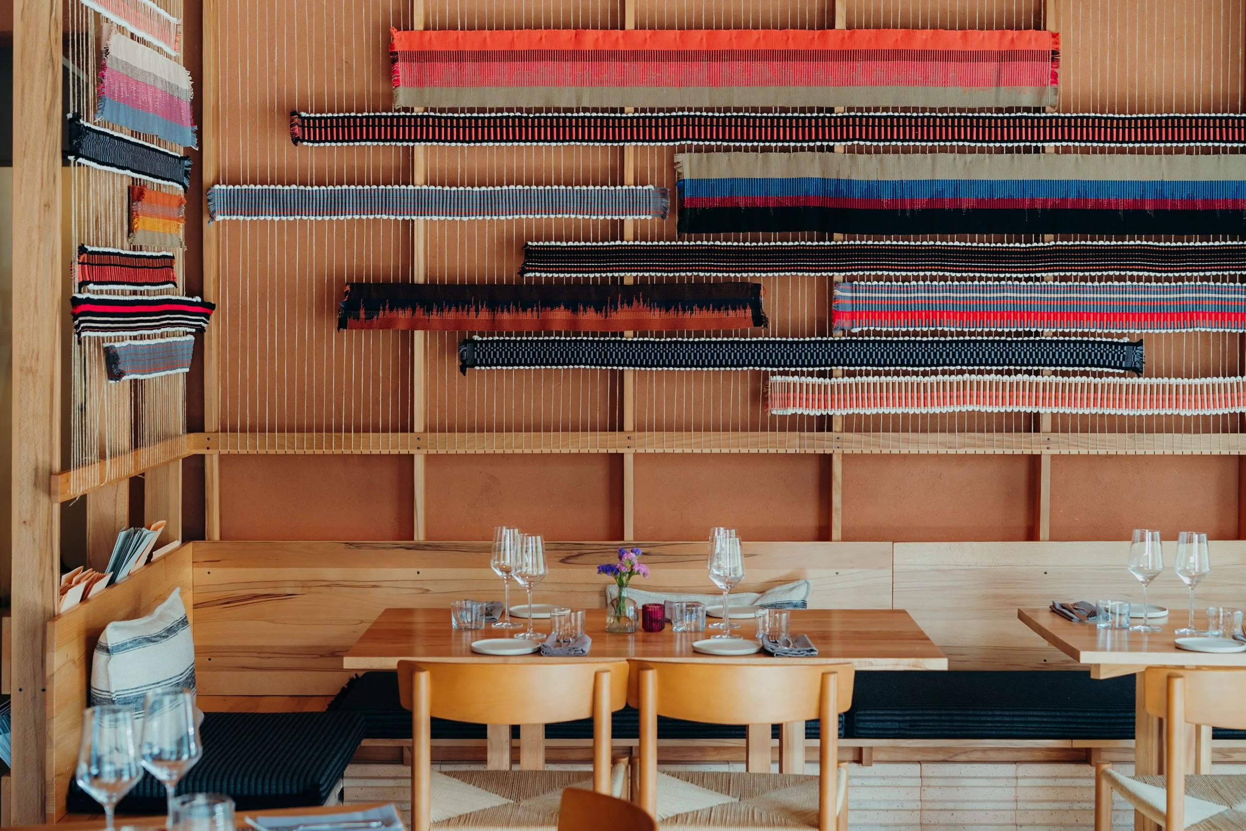 Ein Restaurant mit Holzdekoration, auf der Wand hängen bunte, gewebte Textilien. Im Vordergrund ein gedeckter Tisch mit Gläsern, Tellern und Blumen. An der Seite befinden sich Stühle mit Stoffüberzügen.