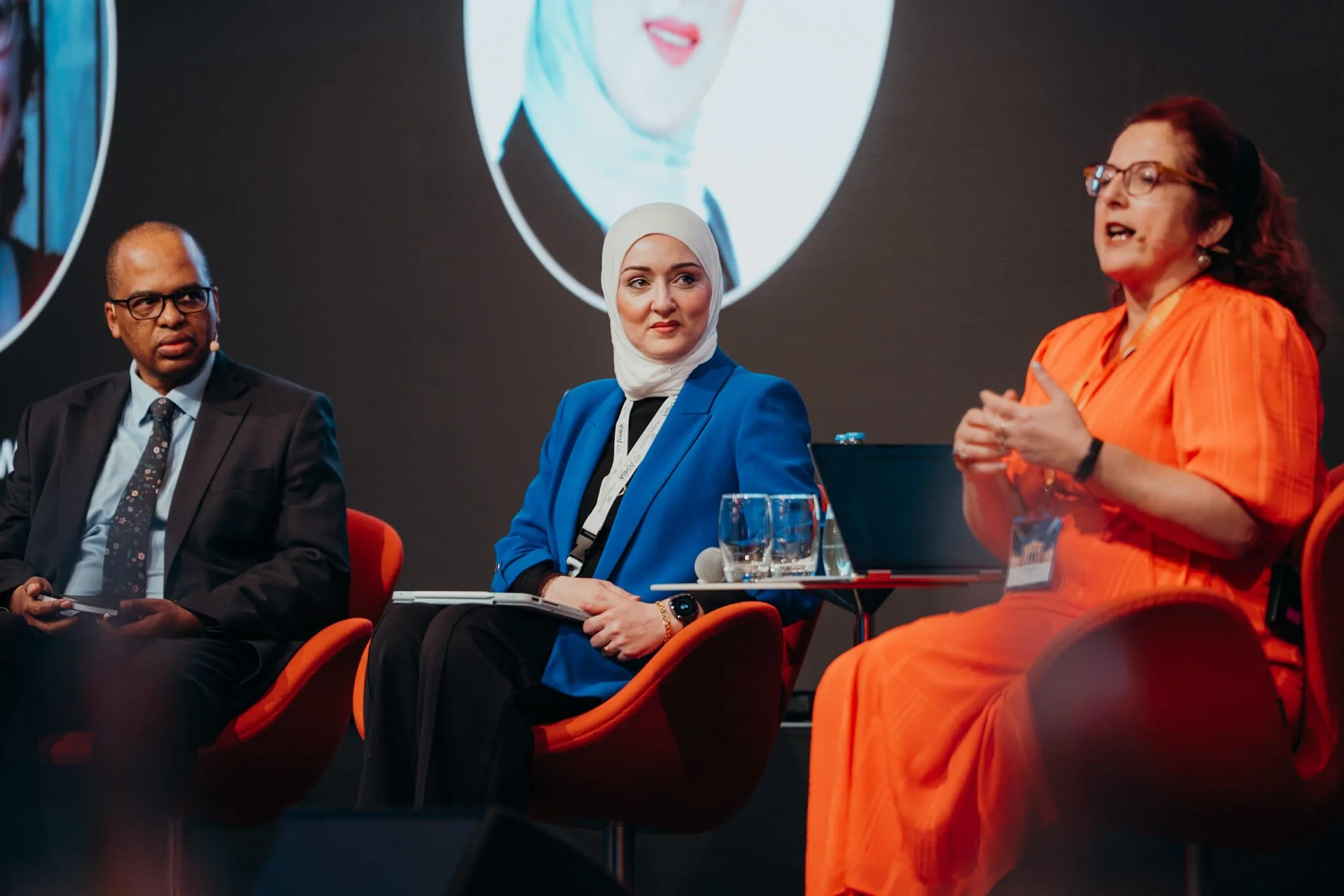 Three diverse women and one man seated on a stage, participating in a panel discussion at a conference. The woman on the right is speaking, wearing an orange dress, and the others are listening attentively. There are large screens behind them display