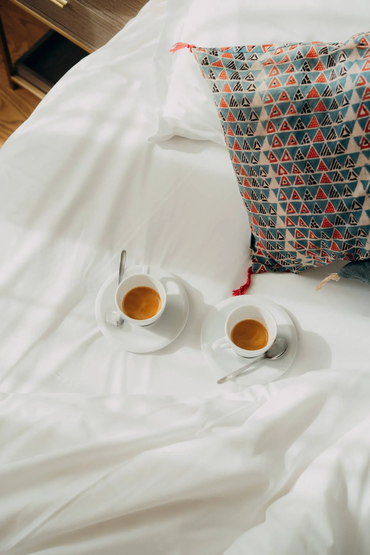Zwei Tassen Kaffee auf weißen Untersetzern mit Löffeln, auf weißem Bettlaken, neben einer Tasche mit bunter Triangel-Muster